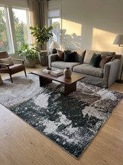 Tapis rectangulaire abstrait aux motifs de taches et de marbrures en vert foncé, gris clair et marron, posé sur un parquet dans un salon lumineux