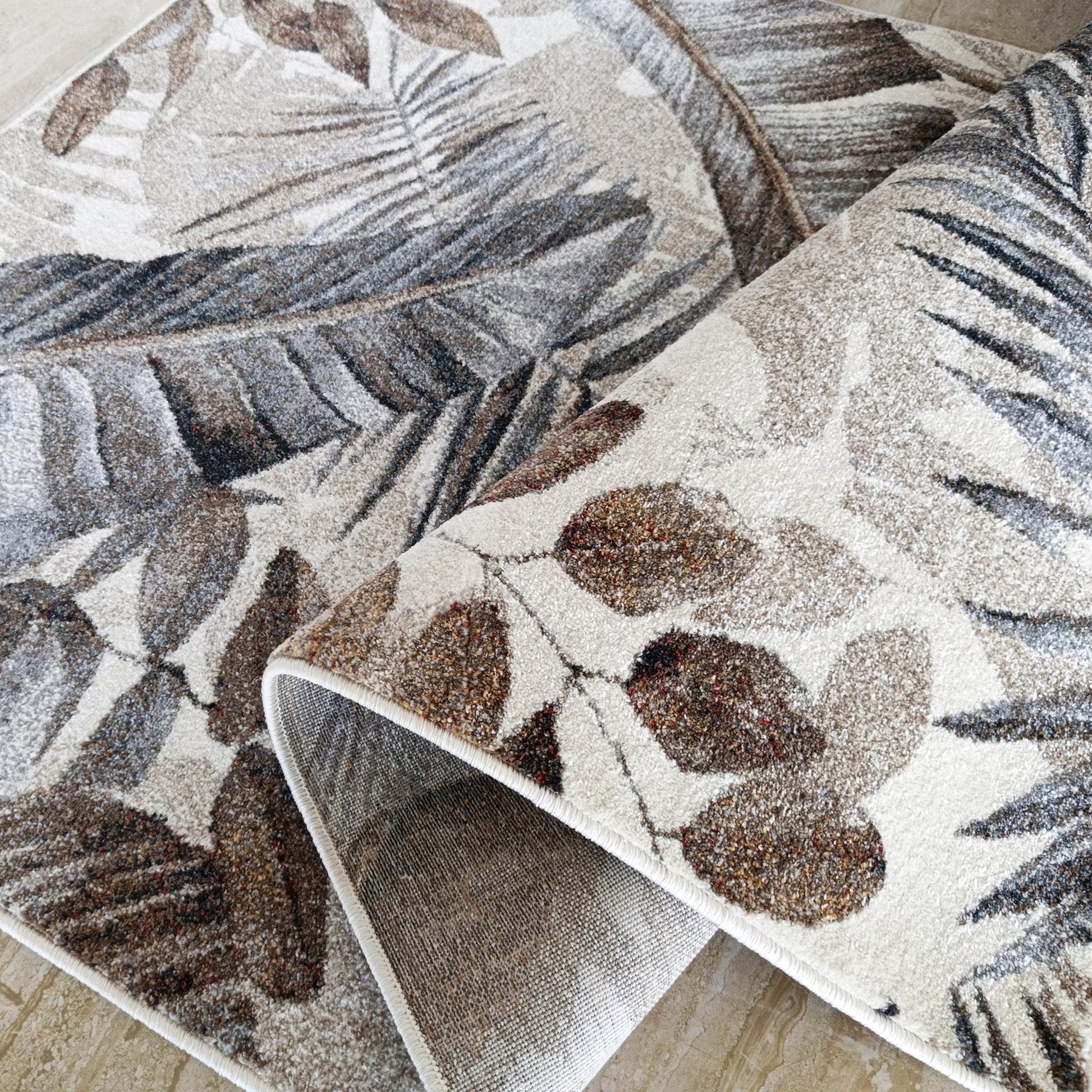 Deux tapis beiges, marron et gris à motifs de feuilles tropicales, partiellement roulés sur un carrelage clair