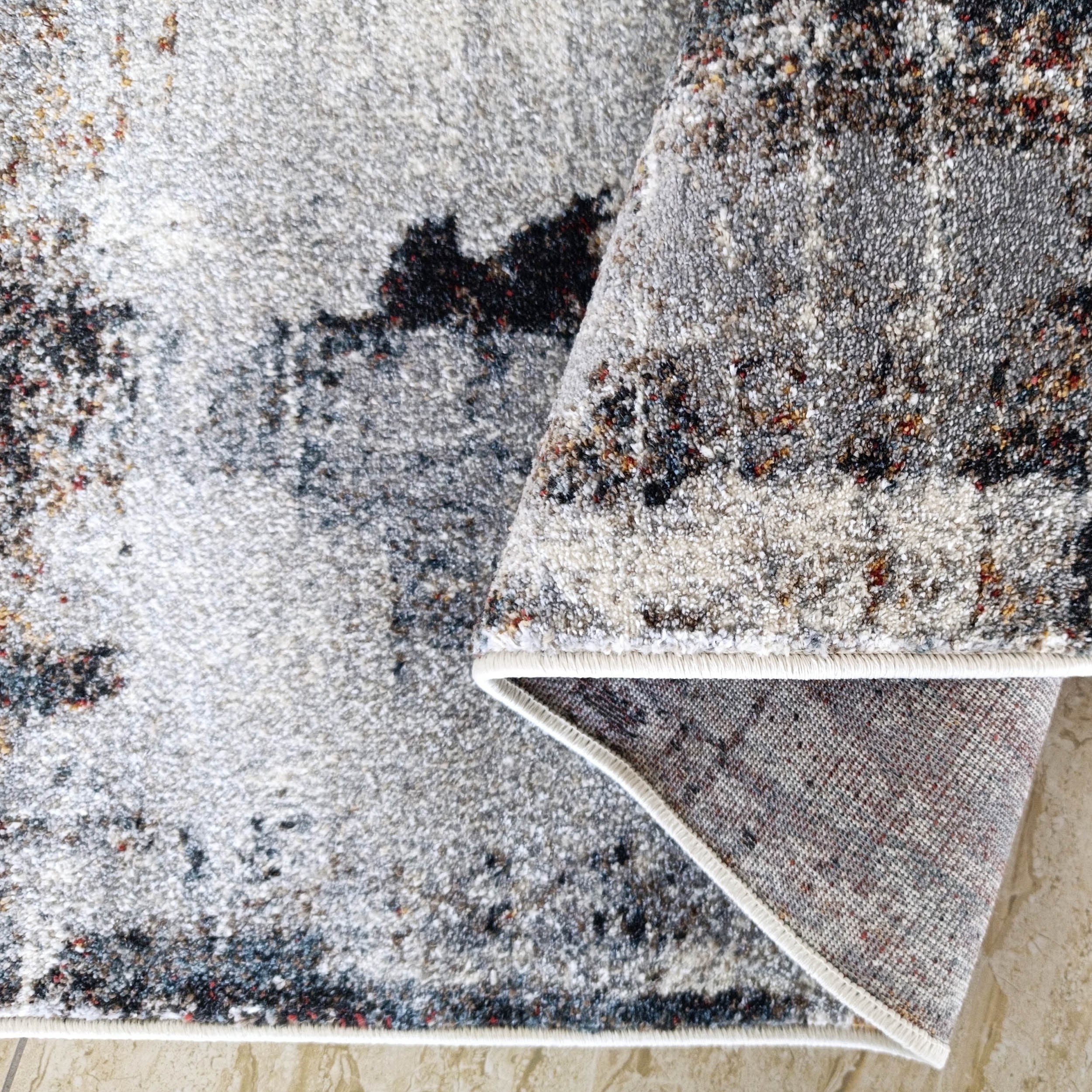 Tapis moelleux à motifs abstraits gris, blancs, noirs et rouges avec bordure blanche, plié sur un carrelage clair veiné
