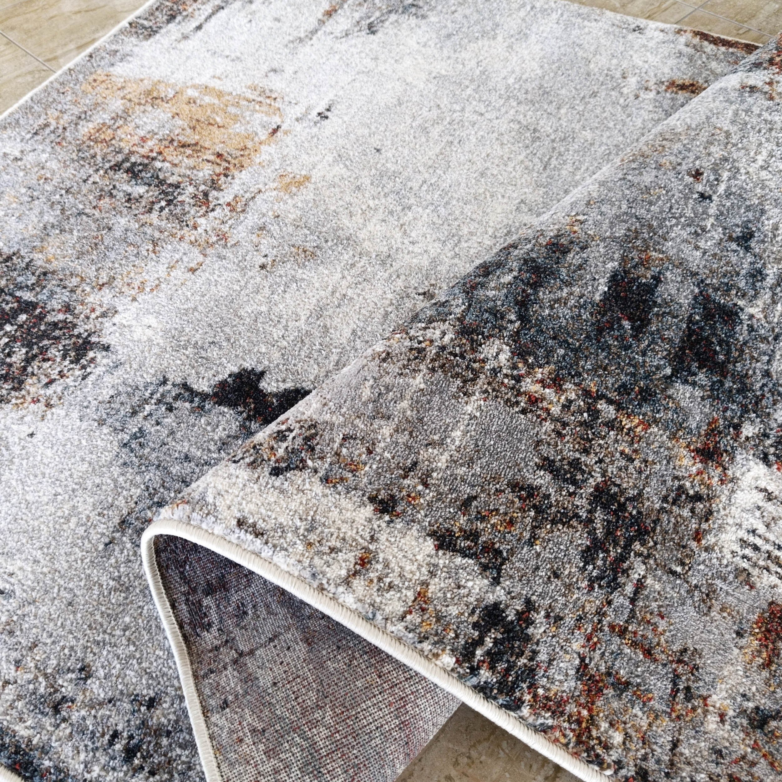Tapis gris moelleux à bordure blanche, motifs abstraits noir, ocre et rouille, roulé sur carrelage