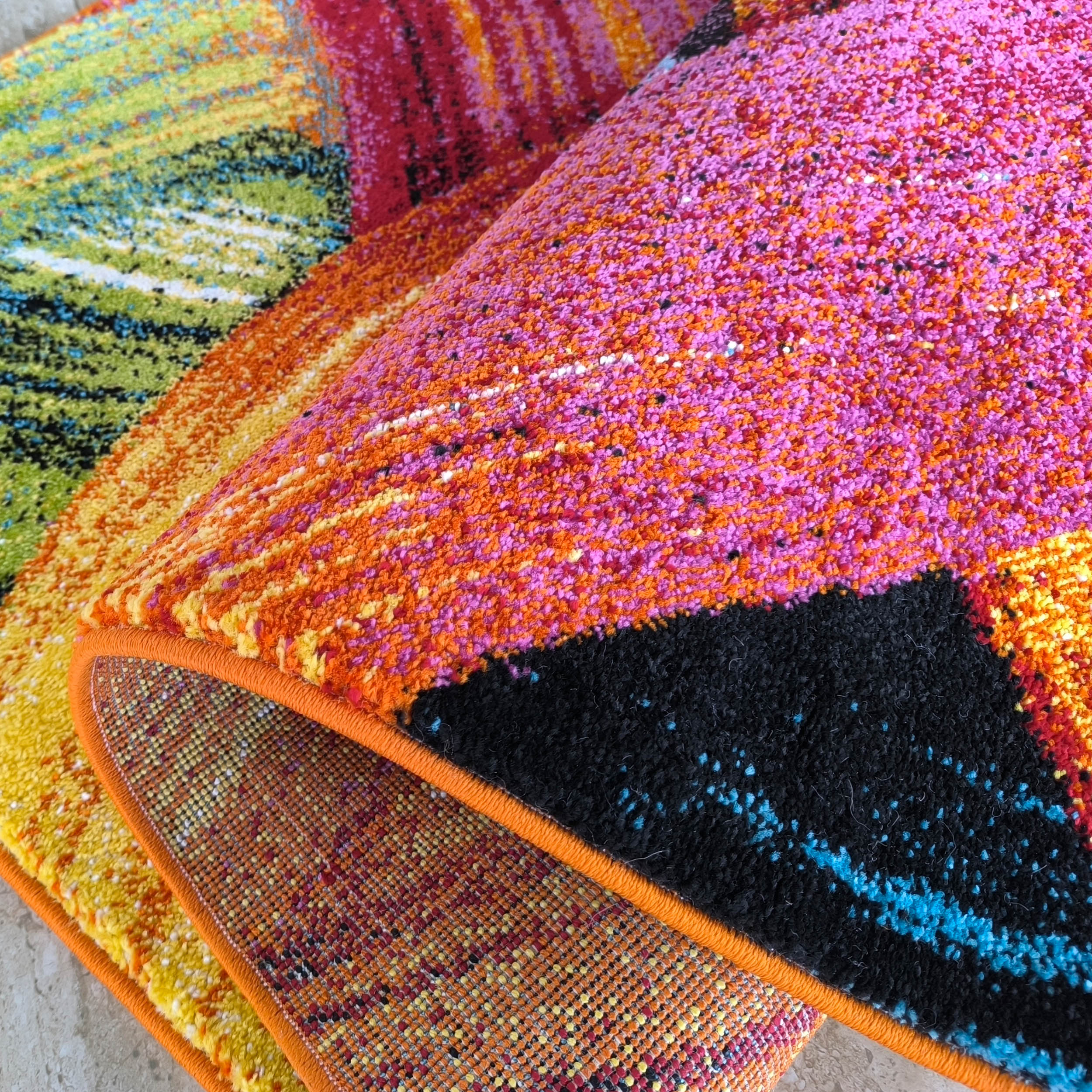 Tapis aux poils épais et vifs motifs abstraits mêlant rose, orange, vert, noir et bleu. Son envers tissé est visible avec un bord surpiqué orange