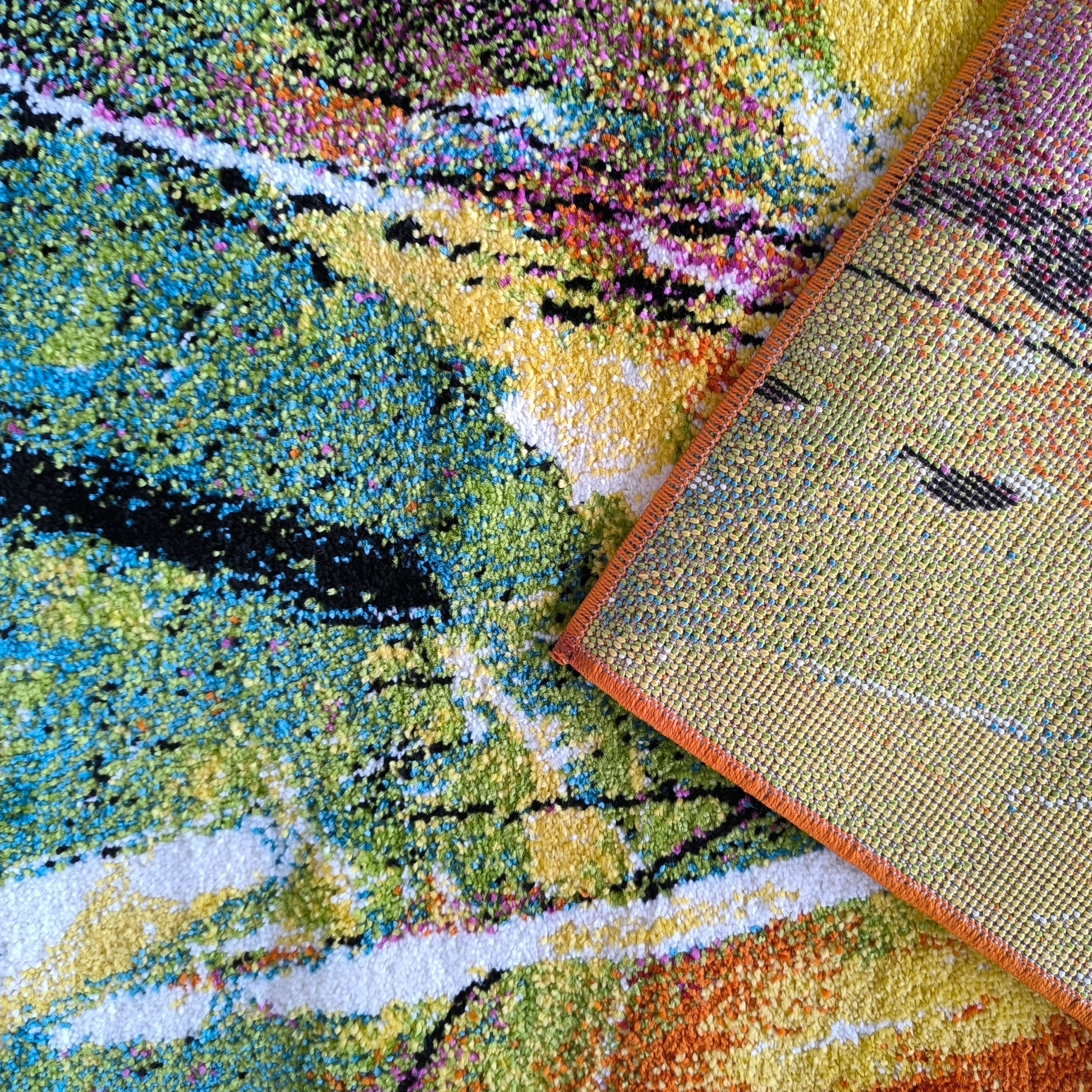 Tapis coloré à texture bouclée avec motifs abstraits verts, bleus, jaunes, oranges, blancs et noirs. Un coin révèle le dos tissé jaune et le bord orange