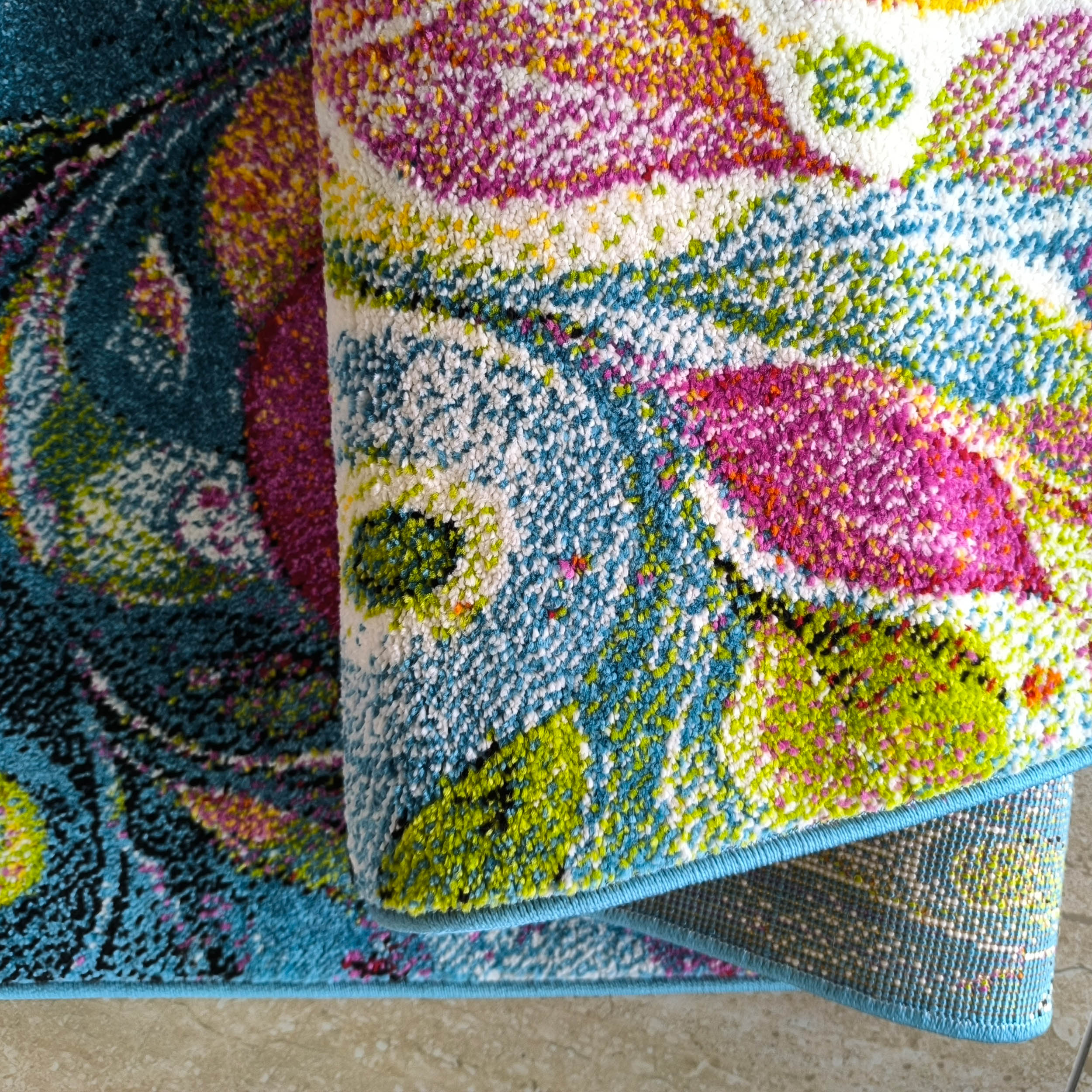 Tapis moelleux aux motifs floraux abstraits très colorés (rose, vert, jaune, bleu, blanc), roulé sur un tapis plus foncé