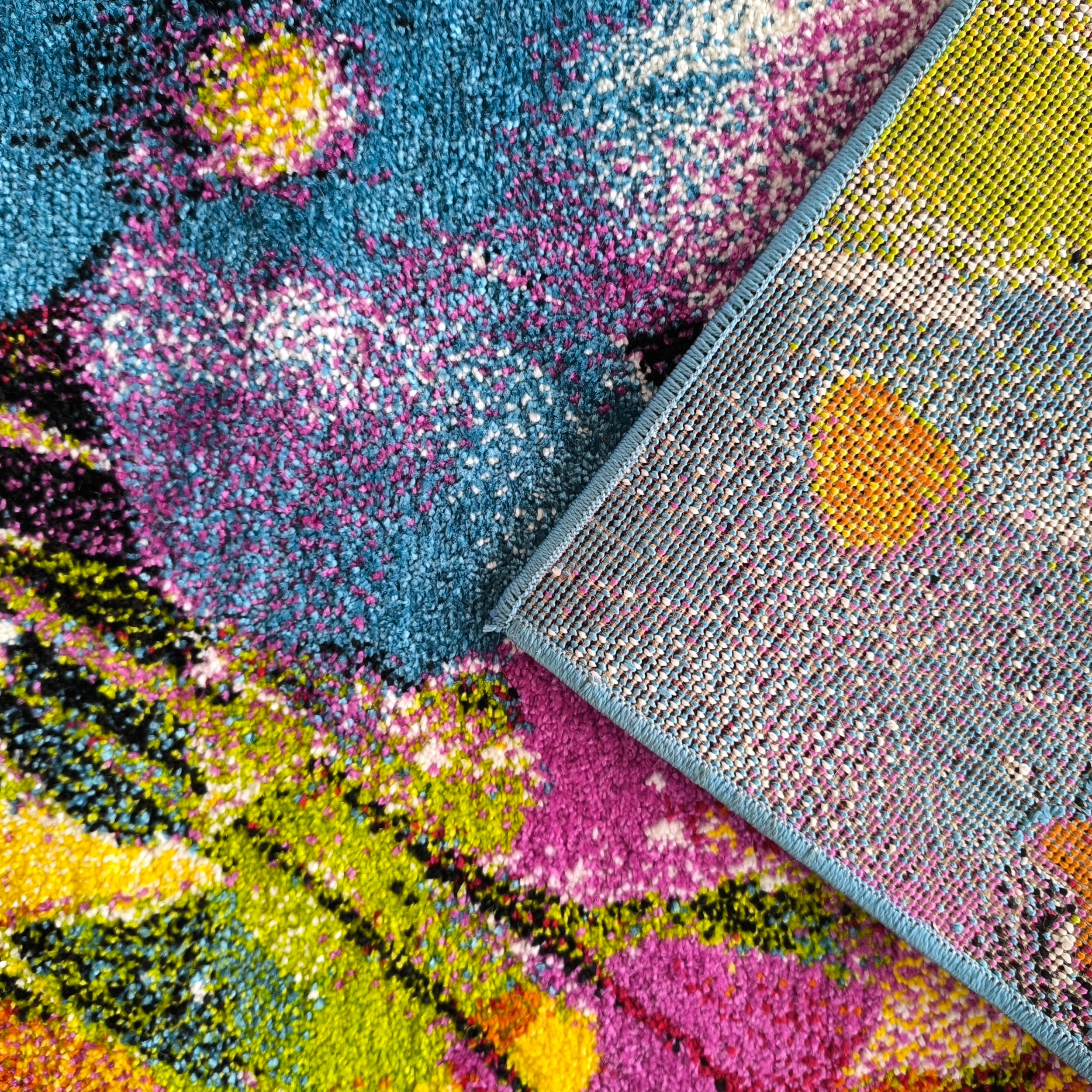 Tapis au poil dense avec des motifs abstraits et mouchetés en bleu, rose, jaune, vert et orange. Un coin est retourné, montrant son envers tissé