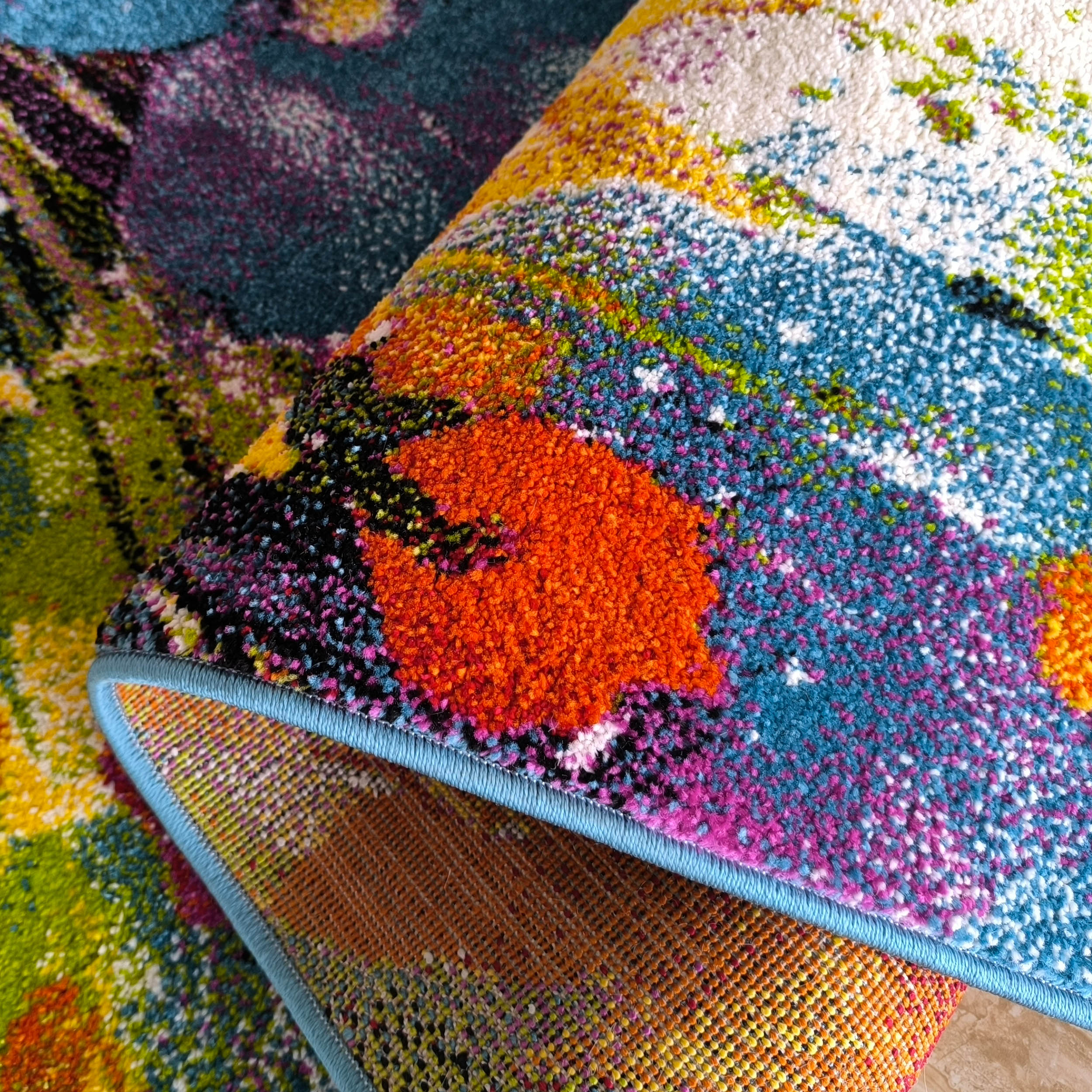 Tapis moelleux et coloré avec des motifs abstraits orange, rose, bleu, vert et blanc. Le bord bleu et le revers tissé sont visibles