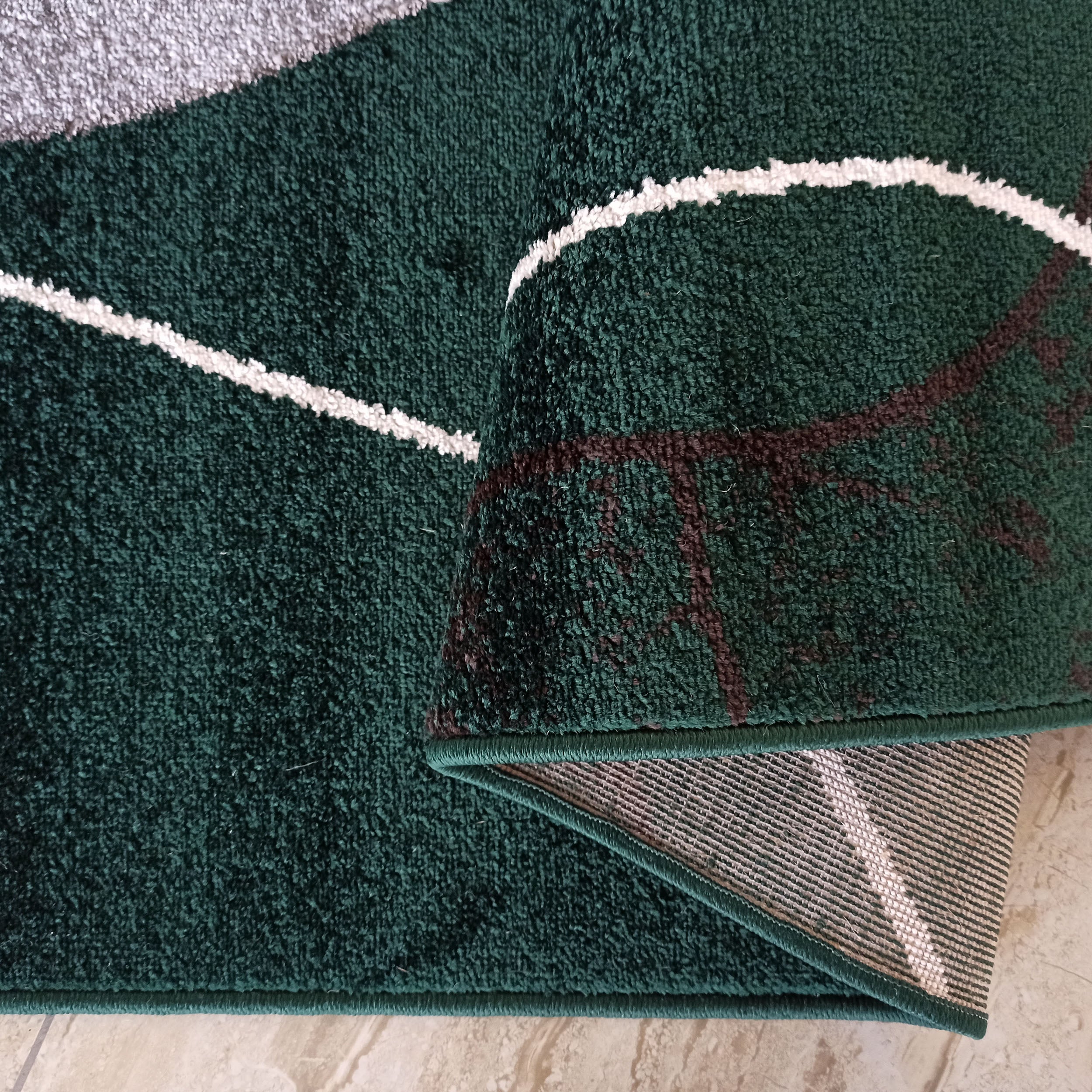 Tapis à poils longs vert foncé avec des motifs abstraits blancs, gris et marron. Il est partiellement roulé, montrant son envers tissé et le carrelage clair en dessous
