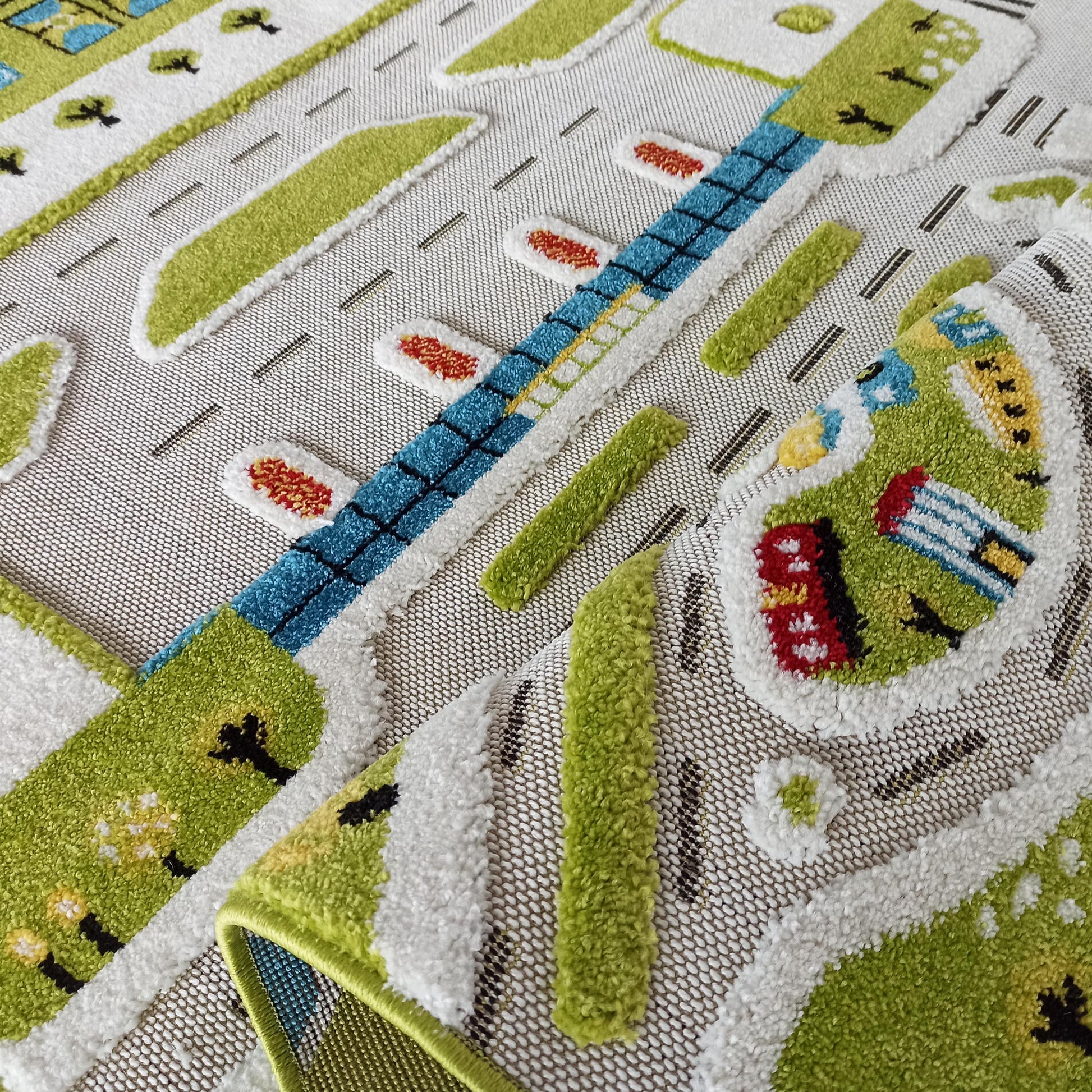 Tapis coloré pour enfant avec un motif de ville en relief: routes grises, herbe verte, bâtiments bleus, rouges et jaunes. Un coin est relevé
