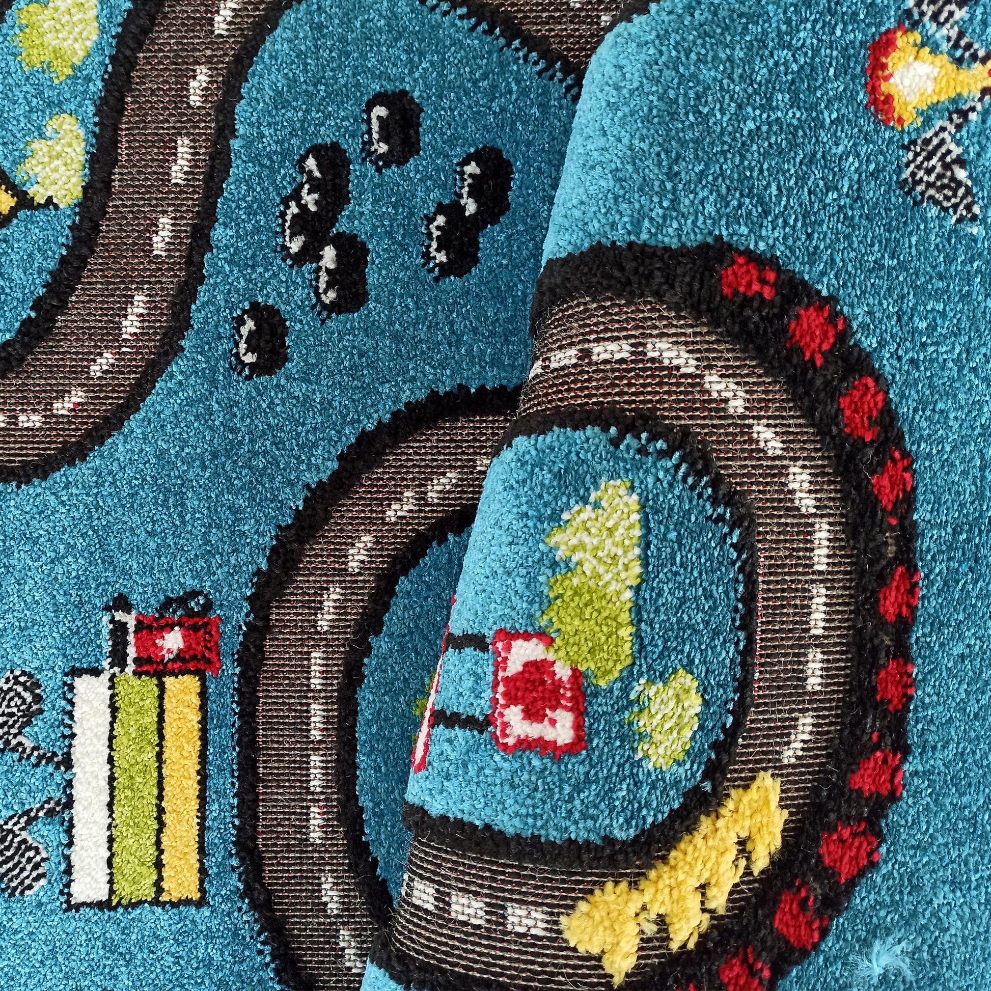 Tapis enfantin turquoise en velours bouclé, partiellement roulé, avec un motif de route, voitures et bâtiments aux couleurs vives: noir, blanc, rouge, jaune et vert