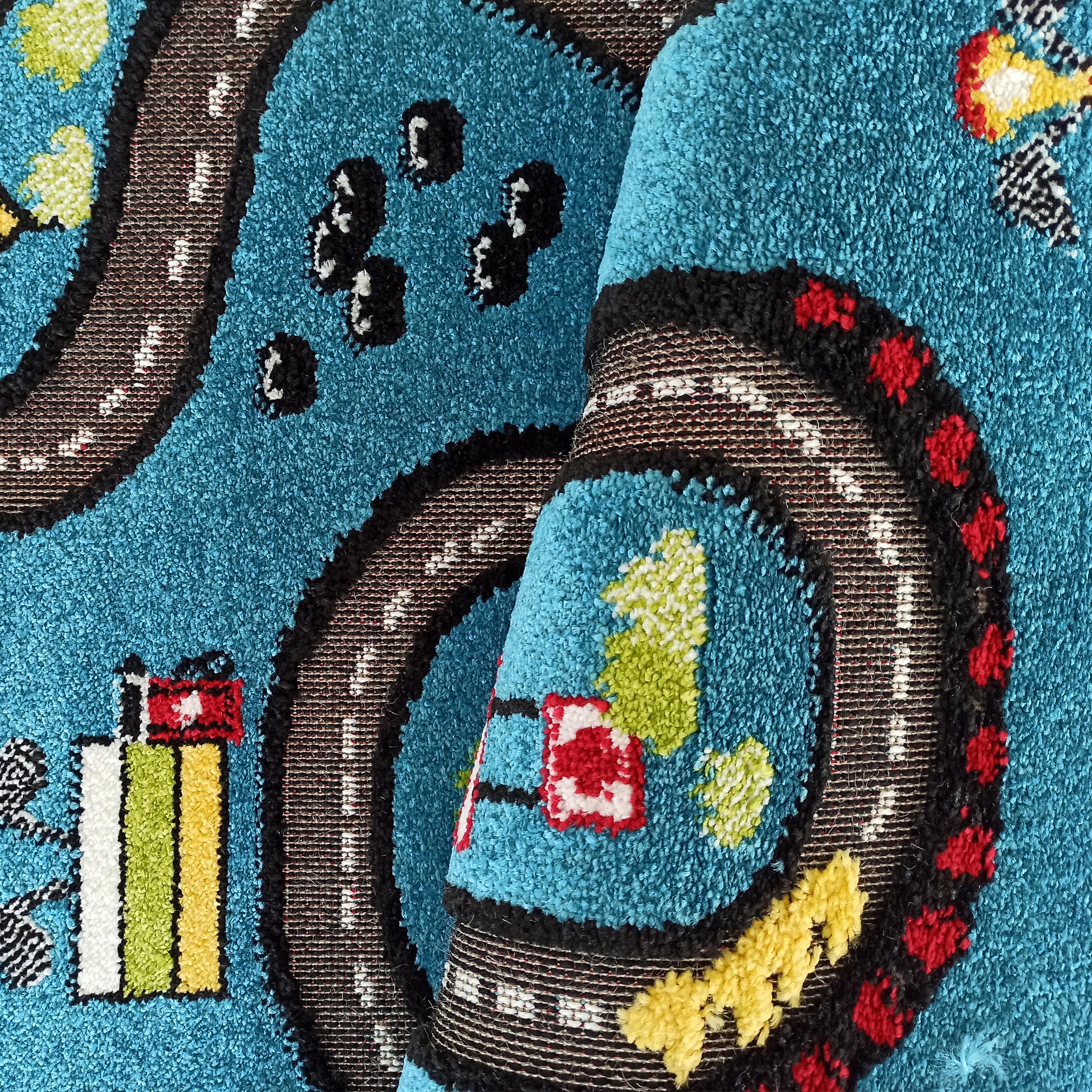 Tapis enfantin turquoise en velours bouclé, partiellement roulé, avec un motif de route, voitures et bâtiments aux couleurs vives: noir, blanc, rouge, jaune et vert