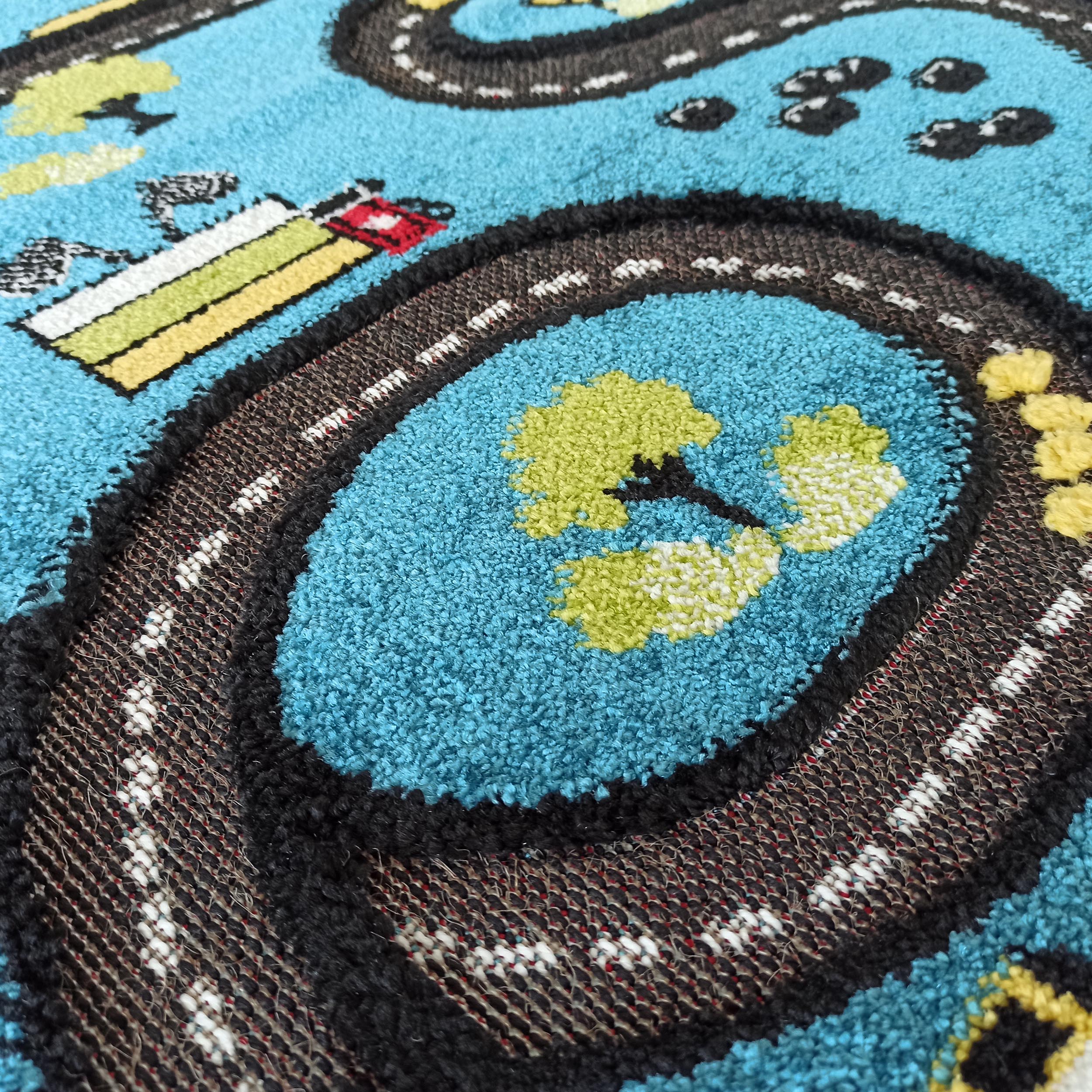 Tapis bleu turquoise texturé avec un motif de routes noires tissées, des marquages blancs, des arbres verts et jaunes, et un bâtiment coloré