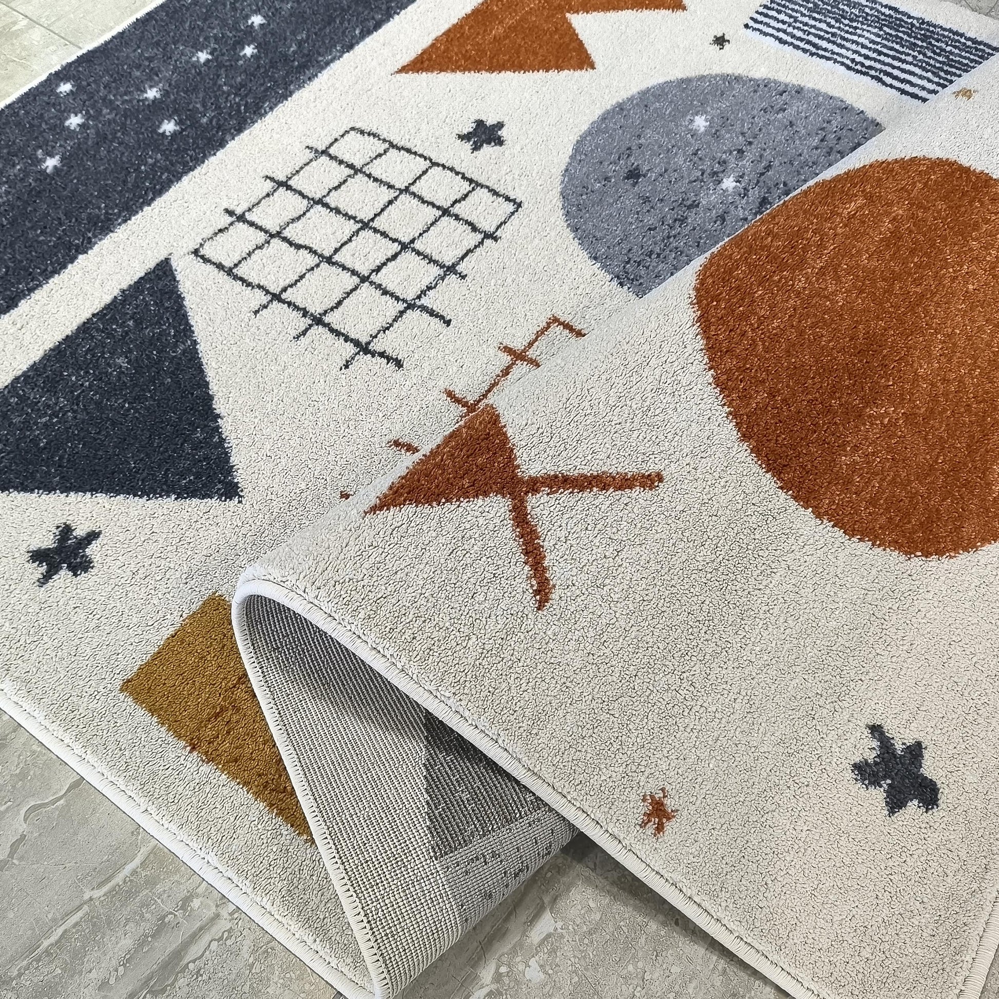 Tapis crème à motifs géométriques gris foncé, orange rouille et noirs, avec le revers visible sur un sol carrelé gris