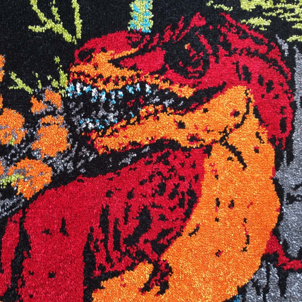 Tapis à la texture veloutée montrant un monstre rouge et orange aux crocs bleus et gris sur fond noir, avec des détails verts