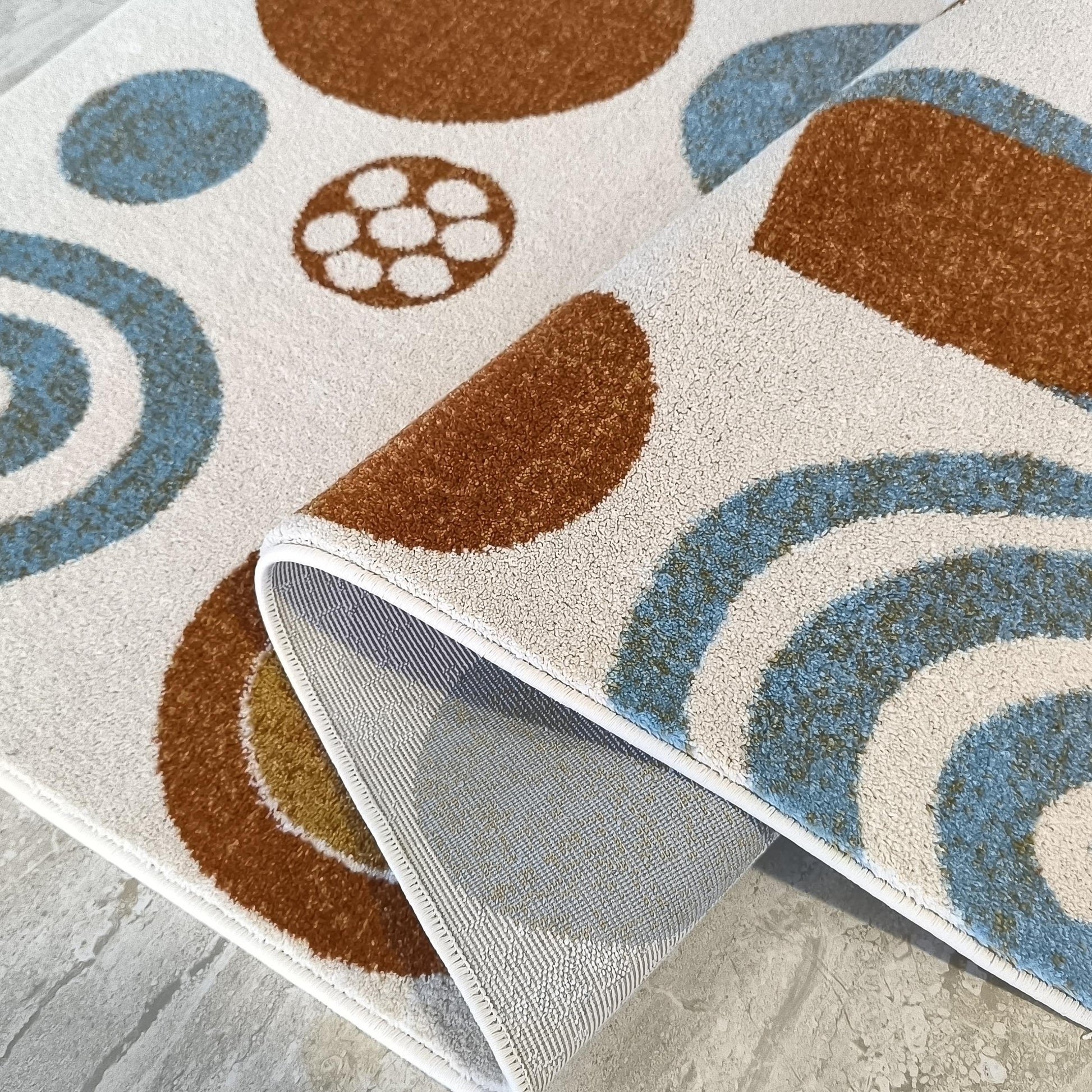 Tapis à motifs géométriques circulaires marron et bleu clair sur fond crème, avec une partie du tapis enroulée montrant son envers texturé gris