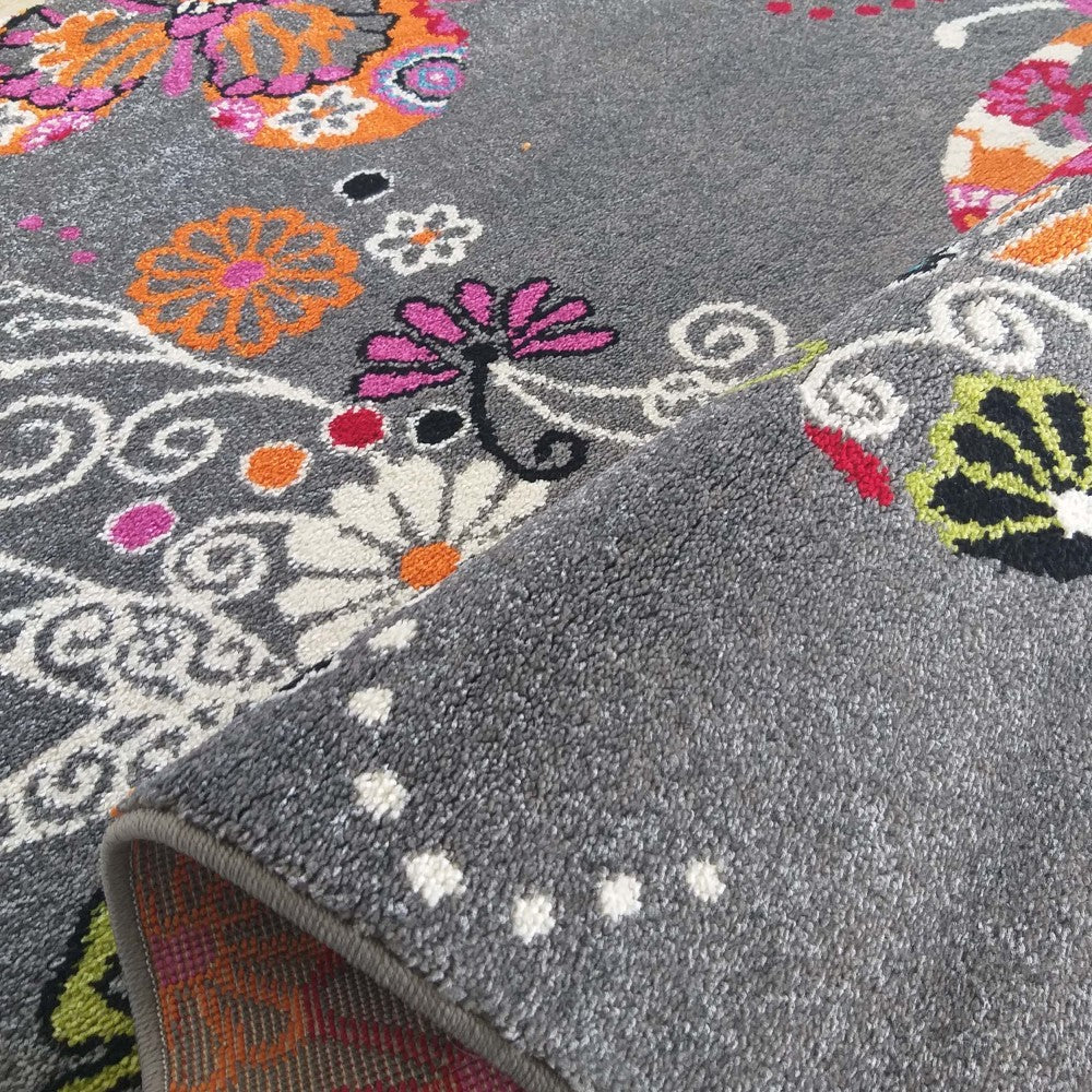 Tapis gris à poils courts avec de vifs motifs floraux et volutes. Le tapis est partiellement roulé, dévoilant son envers gris uni piqué de points blancs