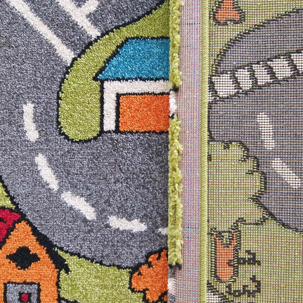 Tapis à motifs de route grise et maisons colorées (orange, bleu, rouge) avec herbe verte. On voit le dessus touffu et l'envers tissé