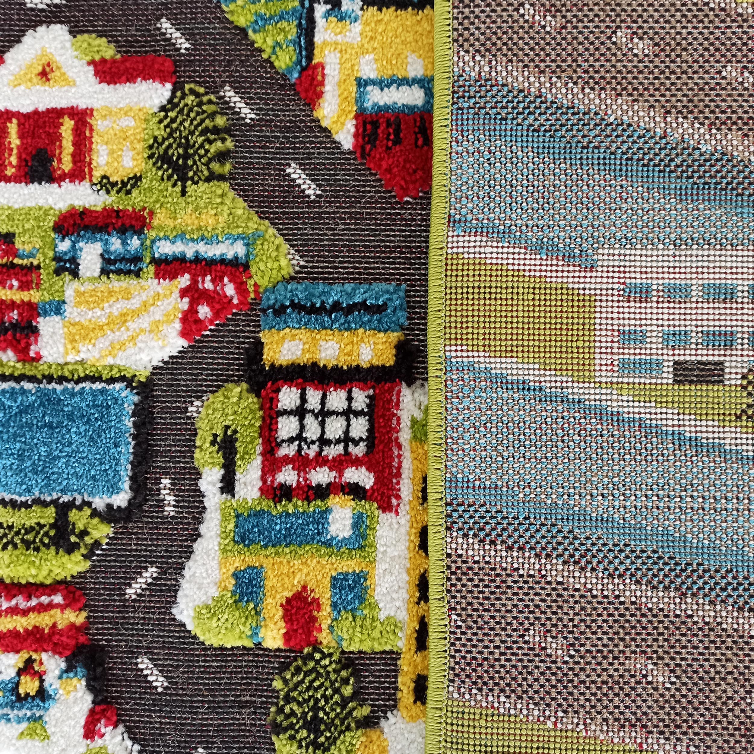 Tapis coloré aux motifs de maisons, arbres et routes sur une surface touffue. Le dos tissé et le bord vert lime sont visibles