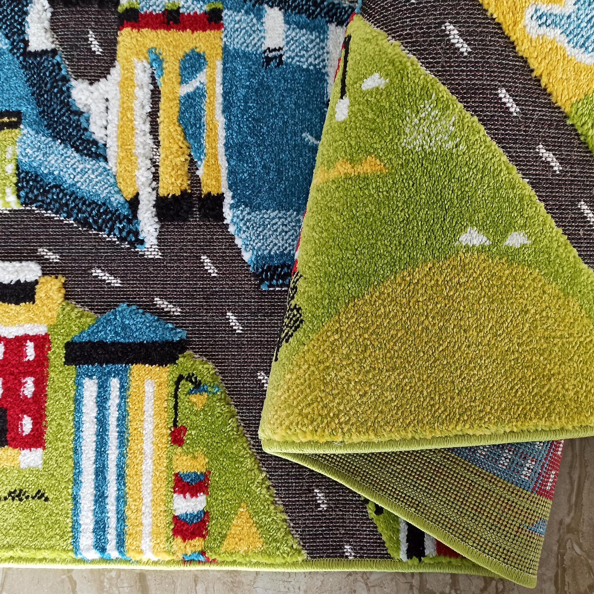 Tapis à motifs urbains enfantins avec routes foncées, bâtiments colorés et véhicules clairs. Sa surface à poils courts est pliée, révélant un envers tissé