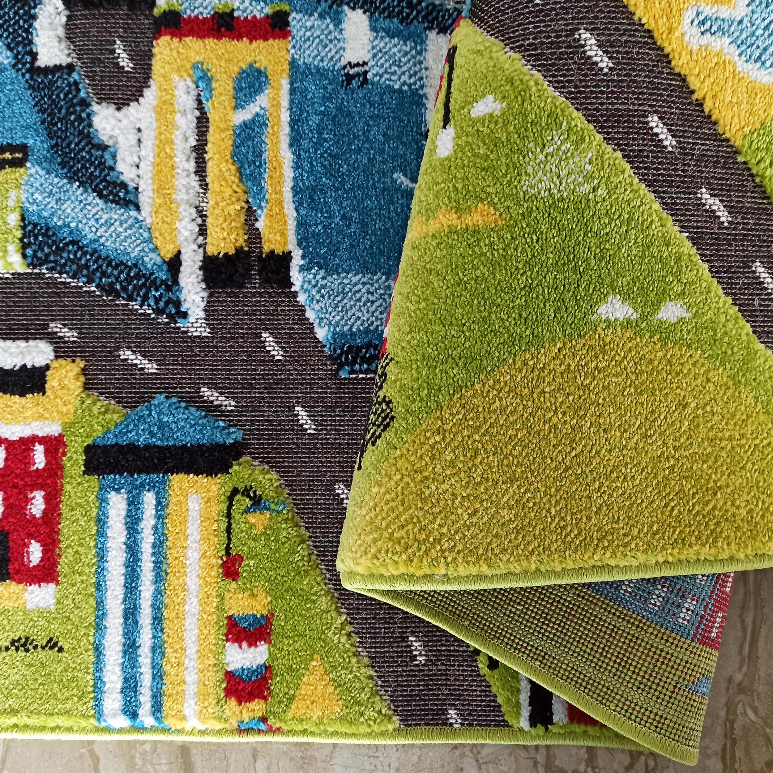 Tapis à motifs urbains enfantins avec routes foncées, bâtiments colorés et véhicules clairs. Sa surface à poils courts est pliée, révélant un envers tissé