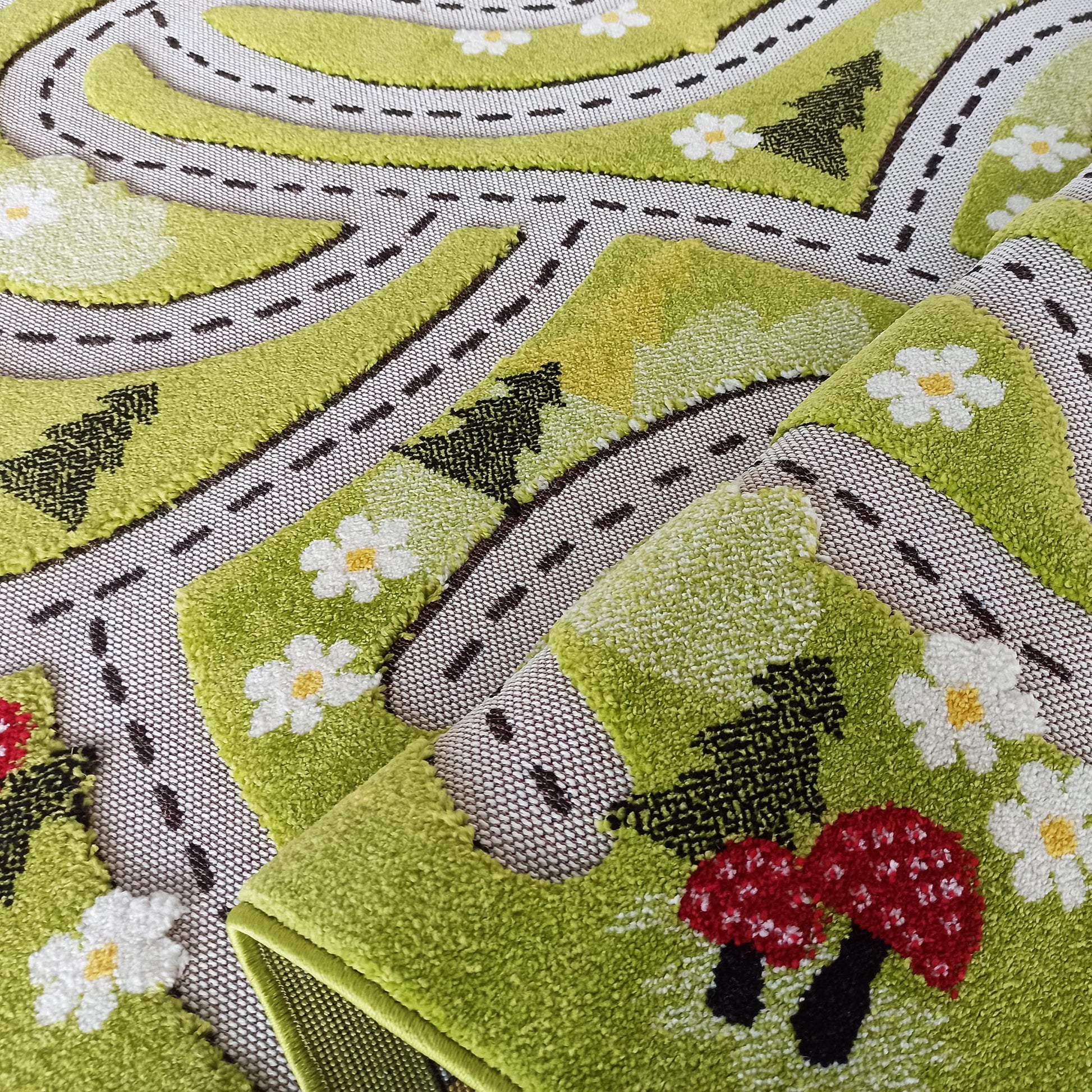 Tapis épais partiellement enroulé représentant un paysage ludique avec des routes, des arbres, des fleurs blanches et des champignons rouges sur un fond vert texturé