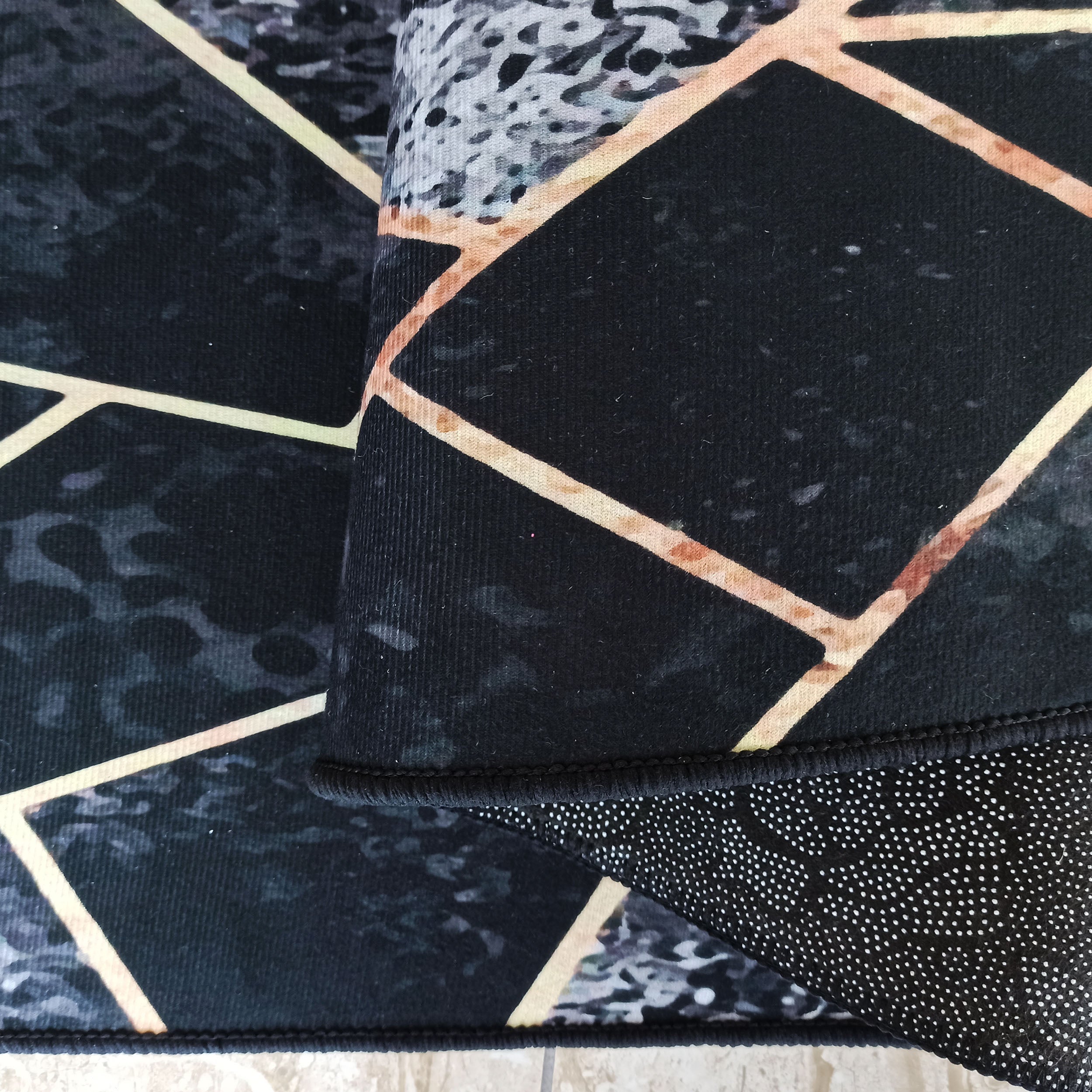 Tapis noir et gris aux motifs géométriques cuivrés et textures marbrées. Le bord et le dessous antidérapant sont apparents