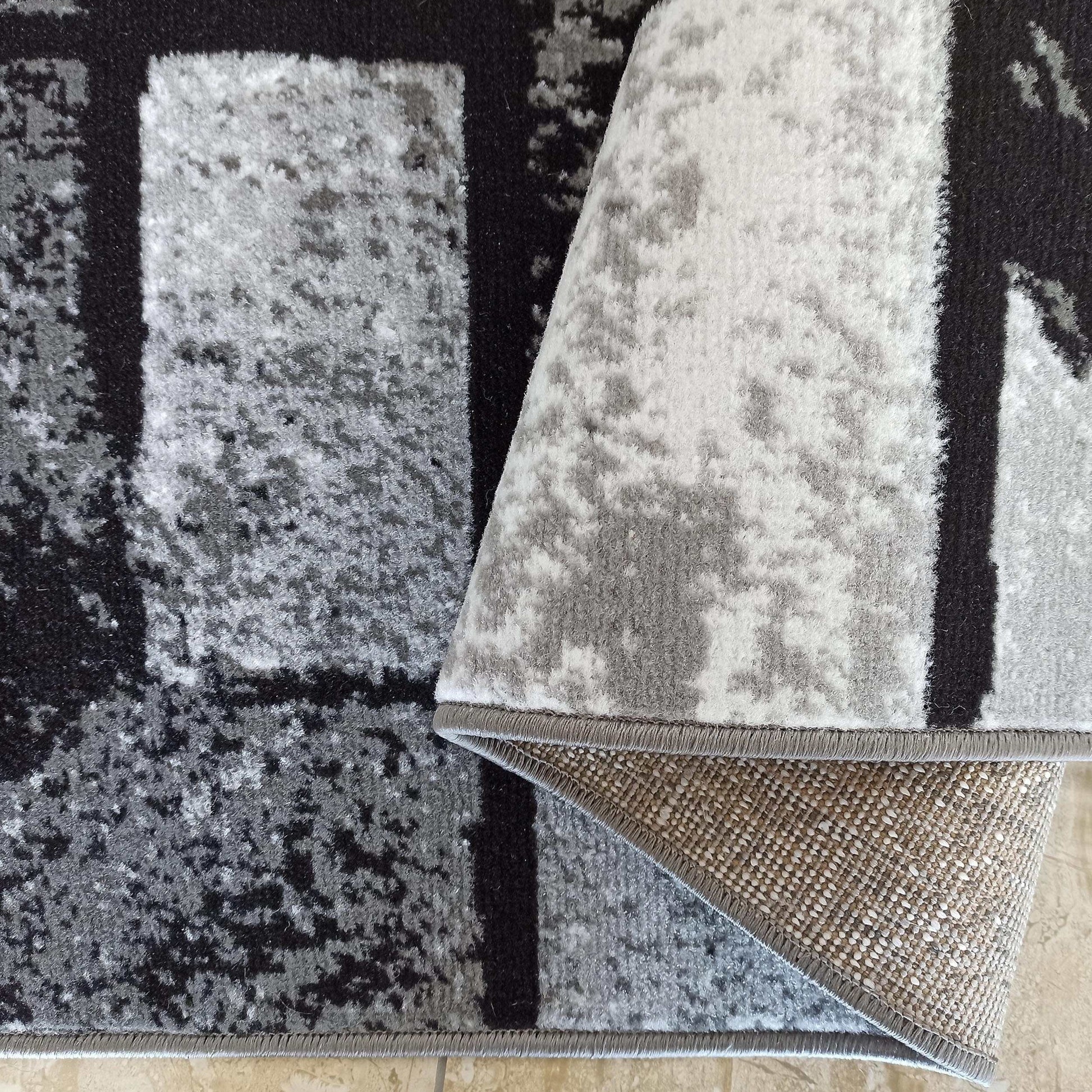 Tapis velouté géométrique gris, noir et blanc, partiellement roulé, envers tissé beige à points blancs, bords gris