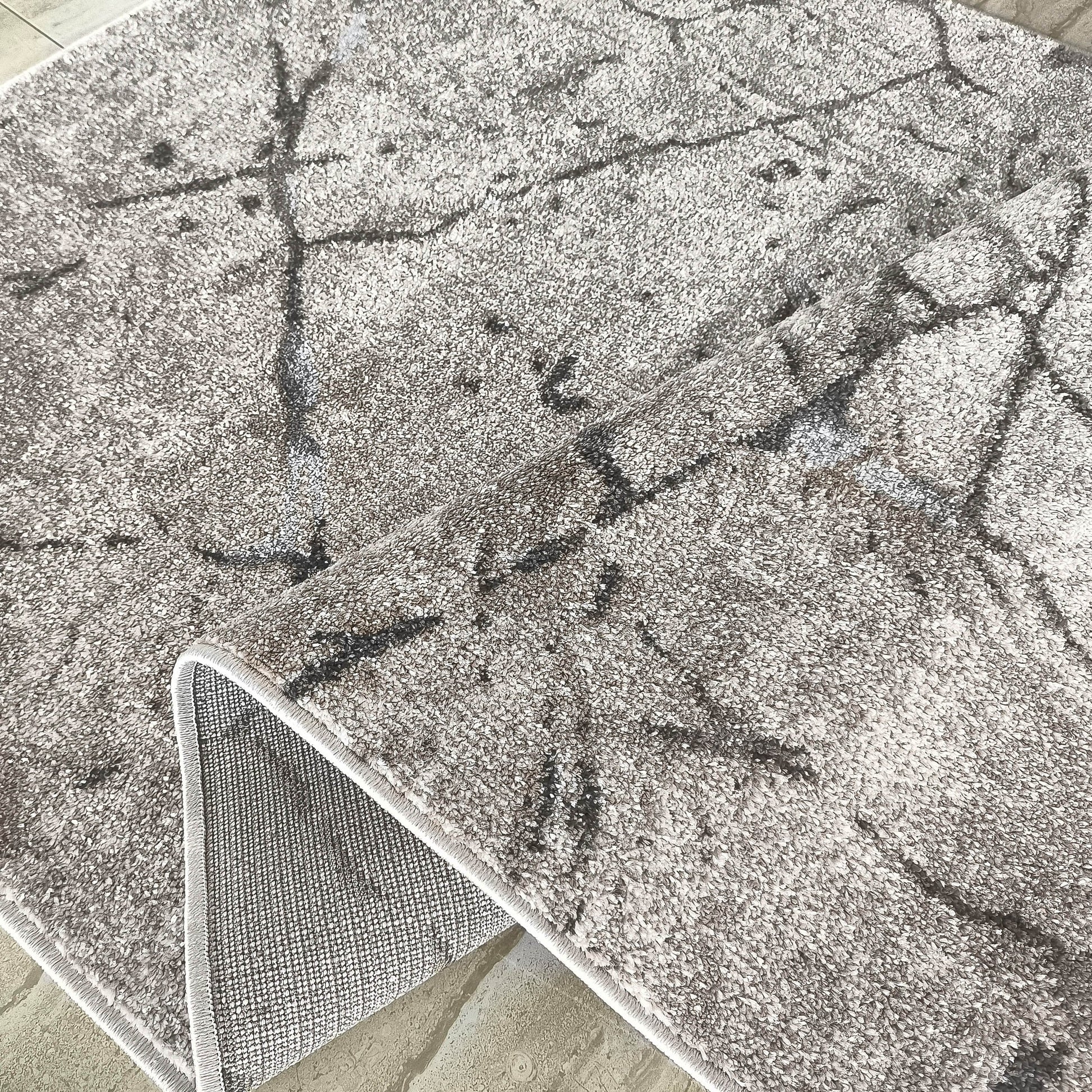 Tapis doux gris clair à motifs abstraits gris foncé, dont un coin replié révèle son envers tissé et le sol carrelé beige