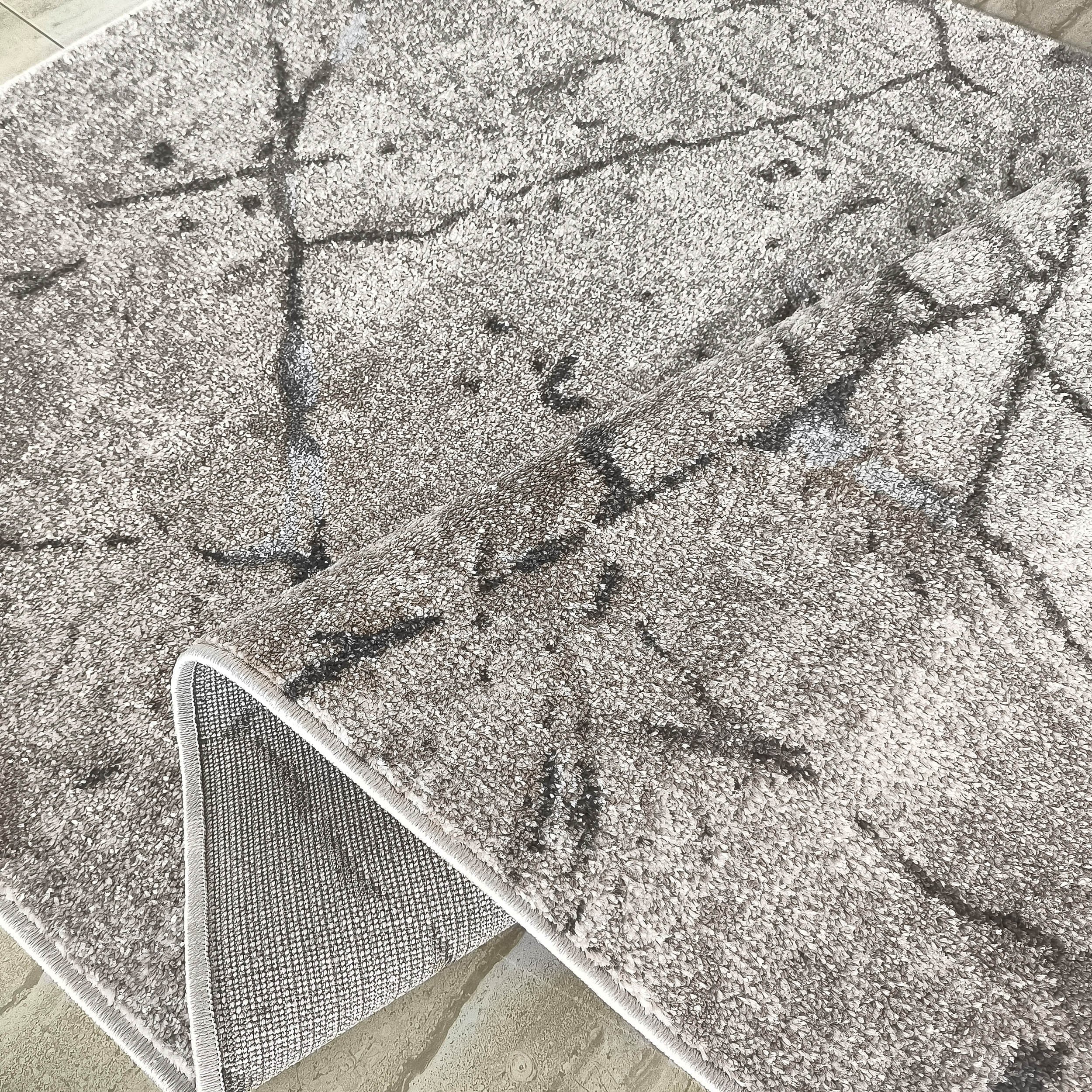 Tapis doux gris clair à motifs abstraits gris foncé, dont un coin replié révèle son envers tissé et le sol carrelé beige