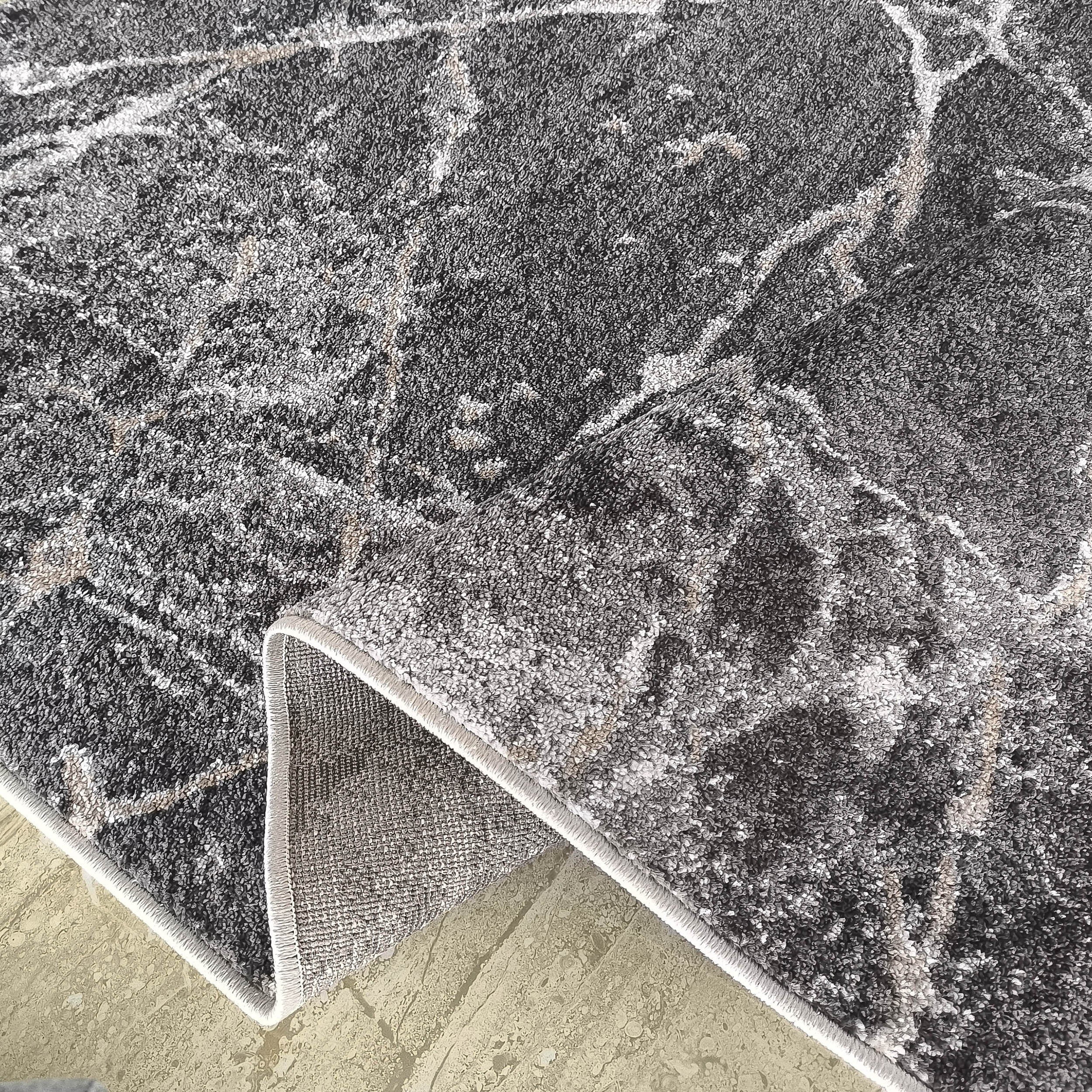 Tapis moelleux gris foncé et clair à motif marbré de veines blanches et beiges, partiellement roulé sur un sol carrelé beige