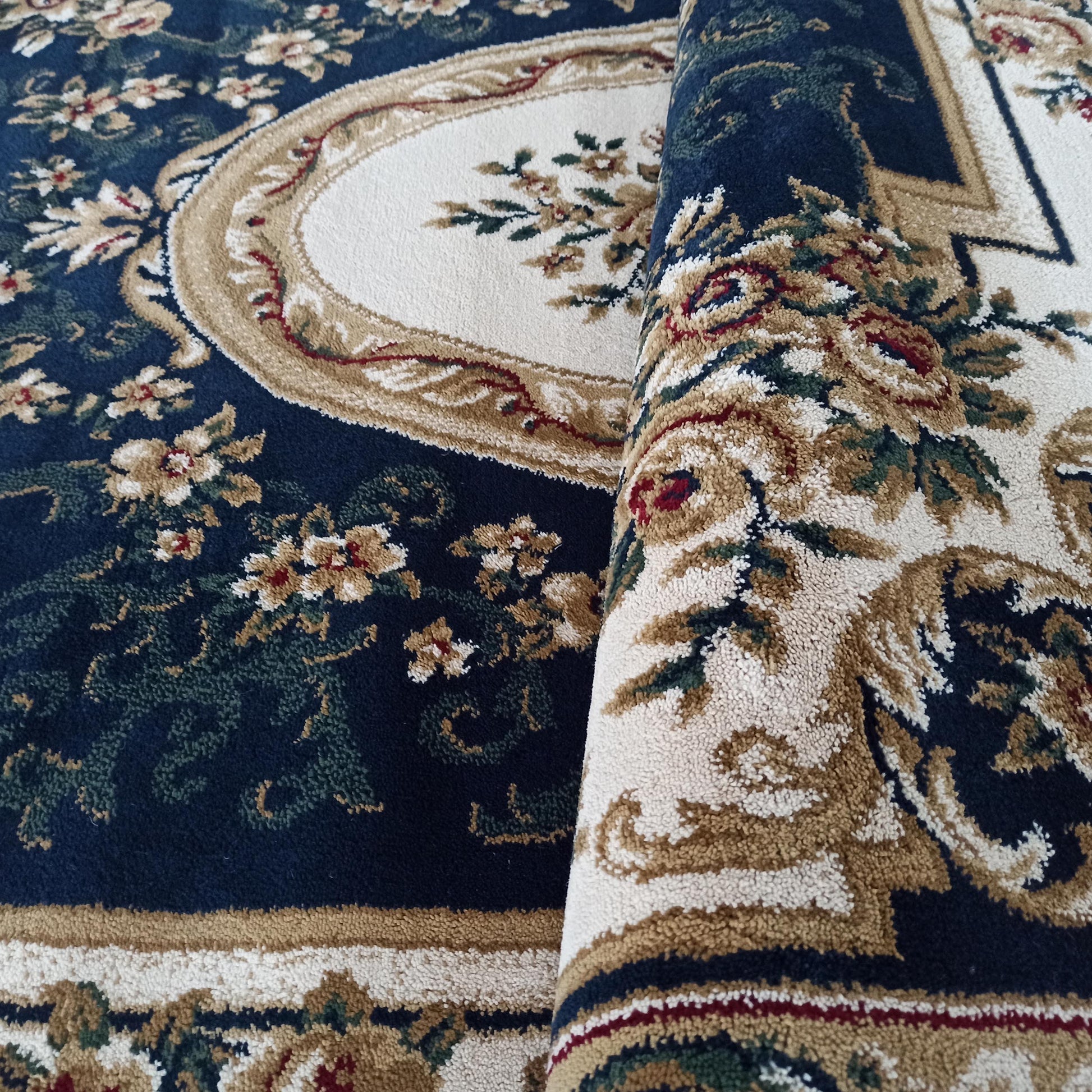 Tapis bleu foncé orné de motifs floraux classiques crème, beige, vert et bordeaux. Une section du tapis est roulée