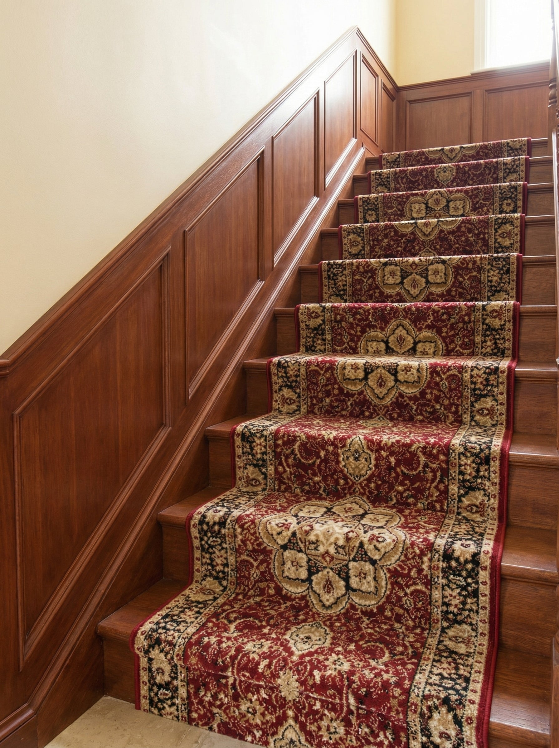 Tapis d'escalier rouge bordeaux à motifs dorés et noirs classiques, posé sur un escalier en bois avec boiseries dans un hall d'entrée