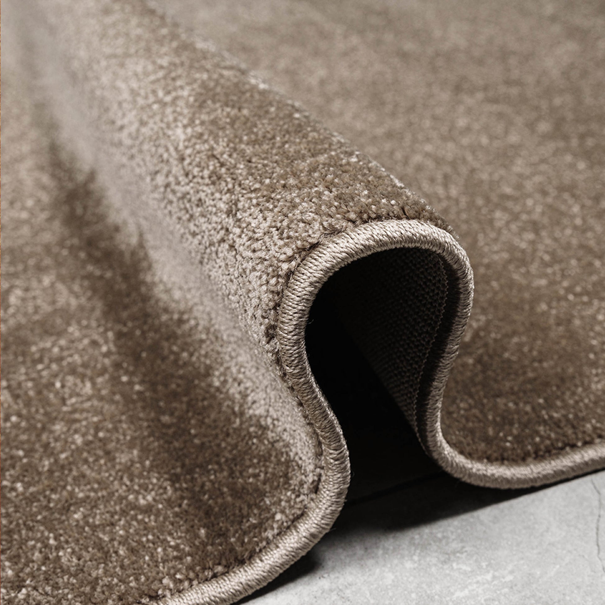 Un tapis marron doux et texturé, dont un côté est roulé pour révéler la finition du bord et le dessous sur un sol gris