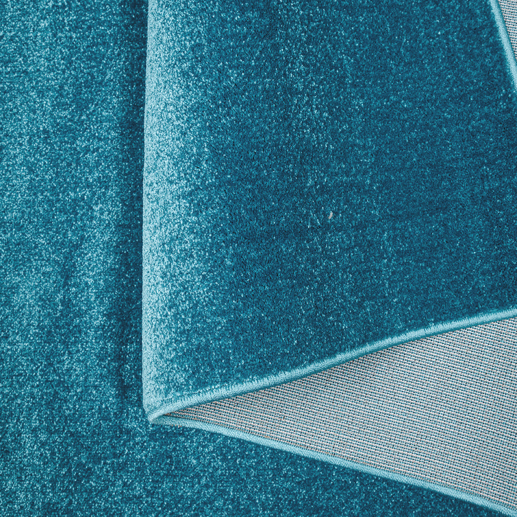 Tapis textile moelleux de couleur bleu-vert, partiellement replié pour révéler son envers gris et sa finition de bordure cousue