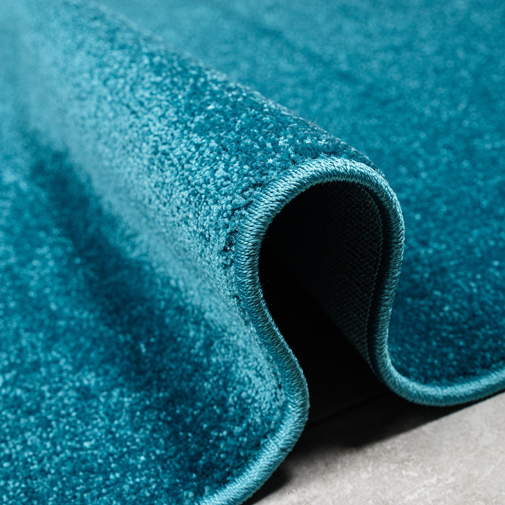 Tapis bleu sarcelle à la texture douce et veloutée, un bord enroulé sur un sol gris
