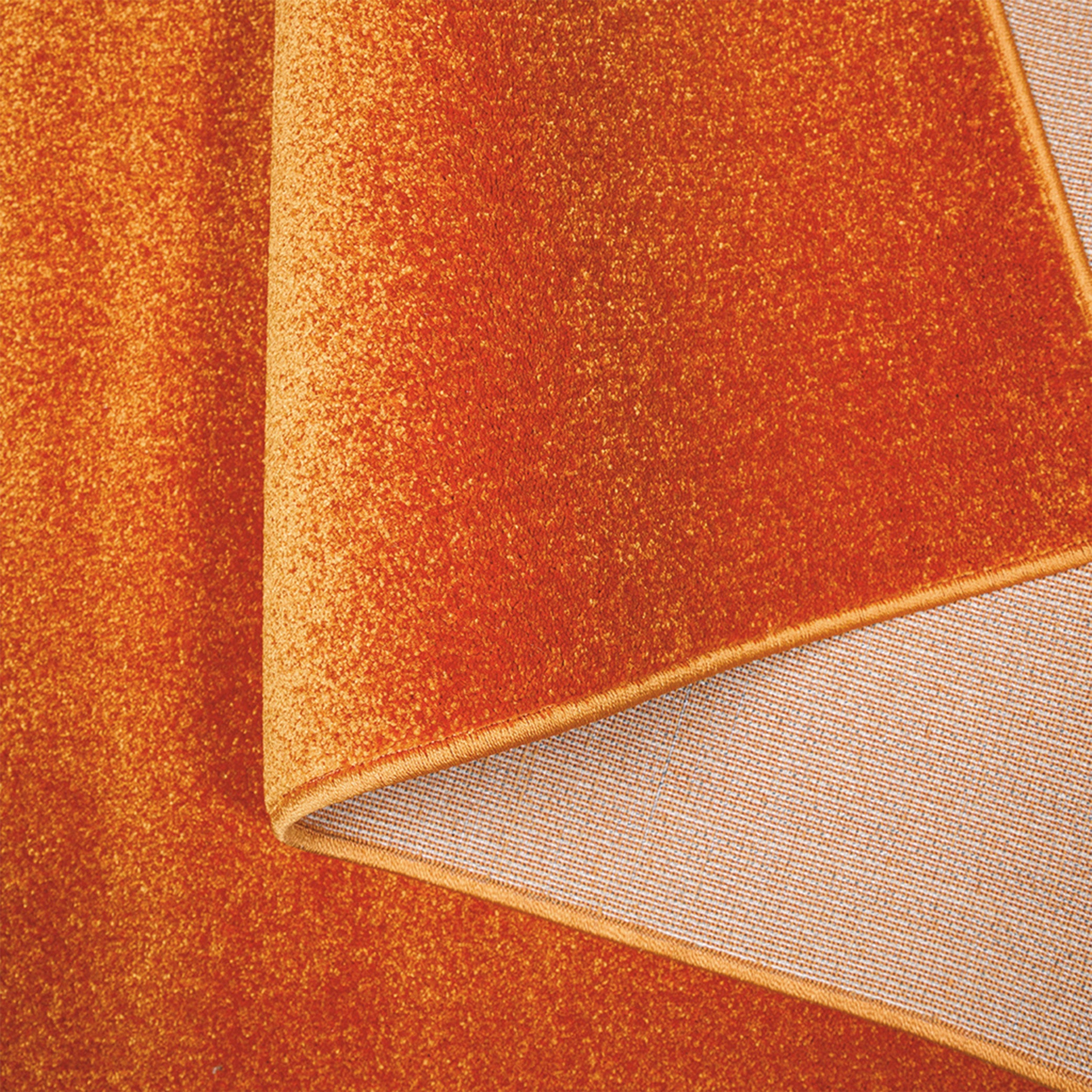 Tapis orange rouille au velours court et texturé, légèrement plié pour exposer son envers tissé de couleur crème à motifs quadrillés