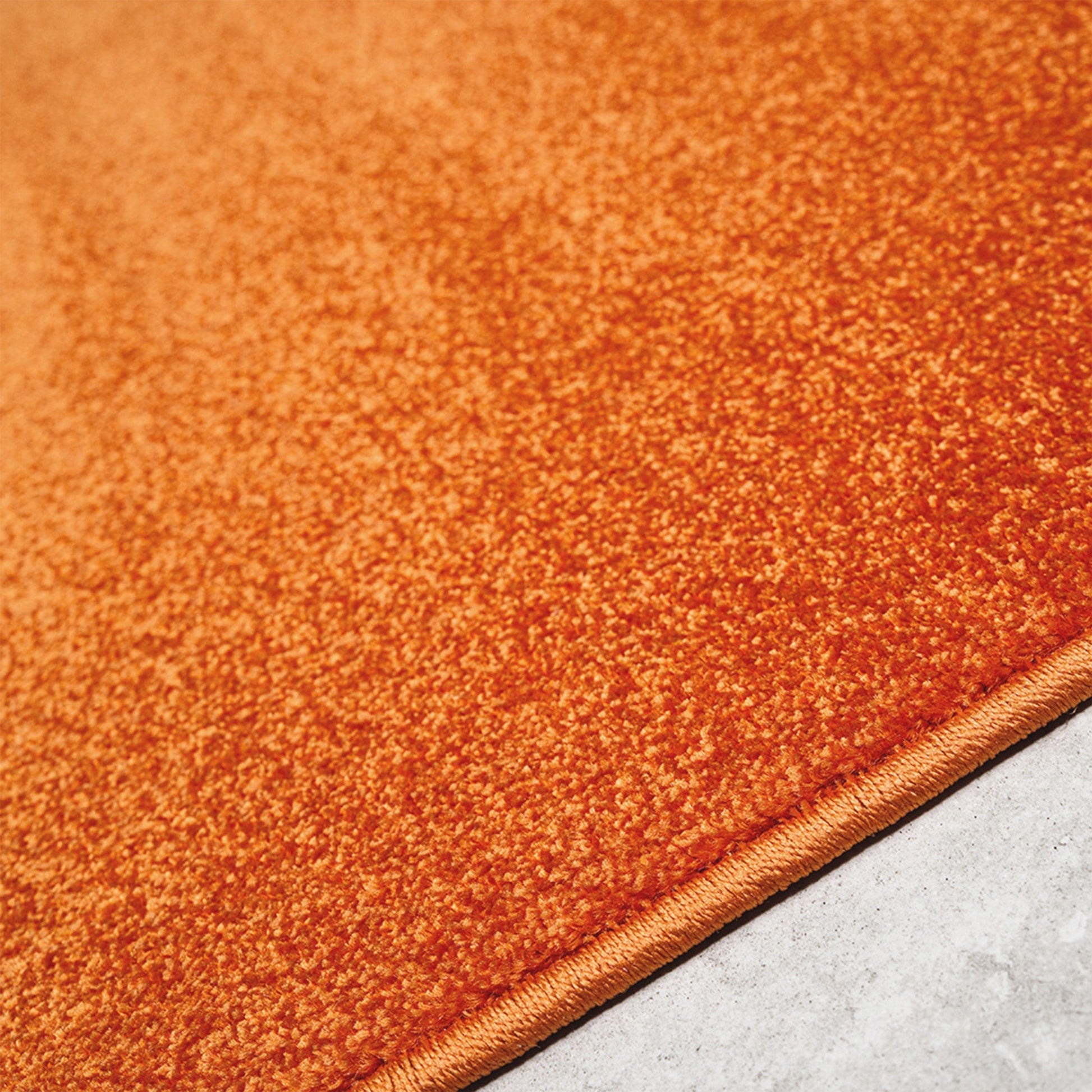Tapis orange vif à texture dense et moelleuse, avec un rebord cousu, posé sur un sol gris uni