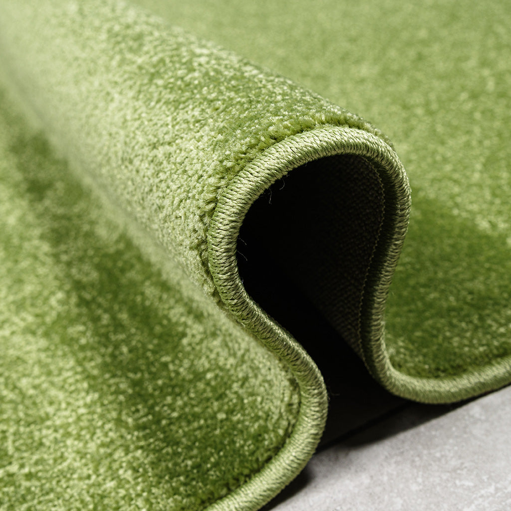 Gros plan sur un tapis vert clair légèrement roulé, montrant sa texture douce et ses bords cousus sur un sol gris