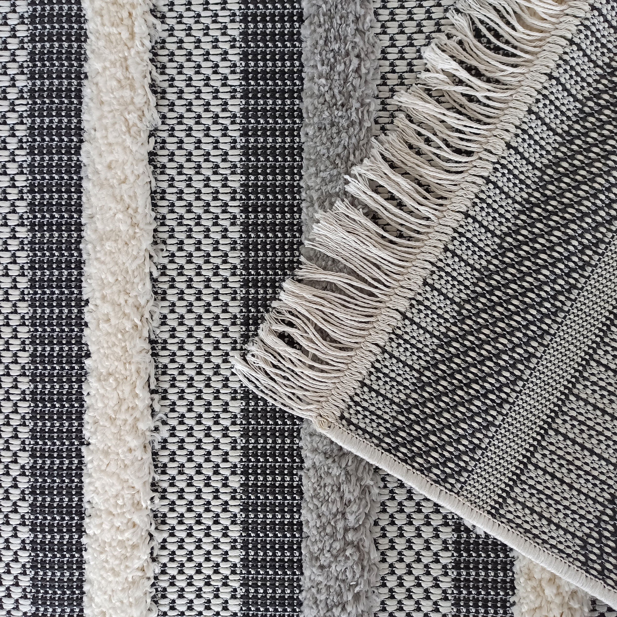 Tapis rayé noir, blanc et gris, composé de bandes à tissage texturé et de bandes à poils longs. Des franges claires sont présentes
