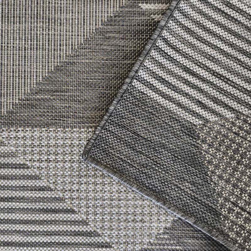 Tapis en textile tissé gris clair et gris foncé, avec des motifs géométriques de rayures et un quadrillage, un coin du tapis est relevé