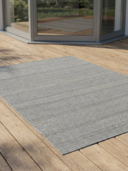 Tapis rectangulaire gris clair tissé, à motifs chevrons et bandes horizontales, posé sur un plancher en bois dans un salon