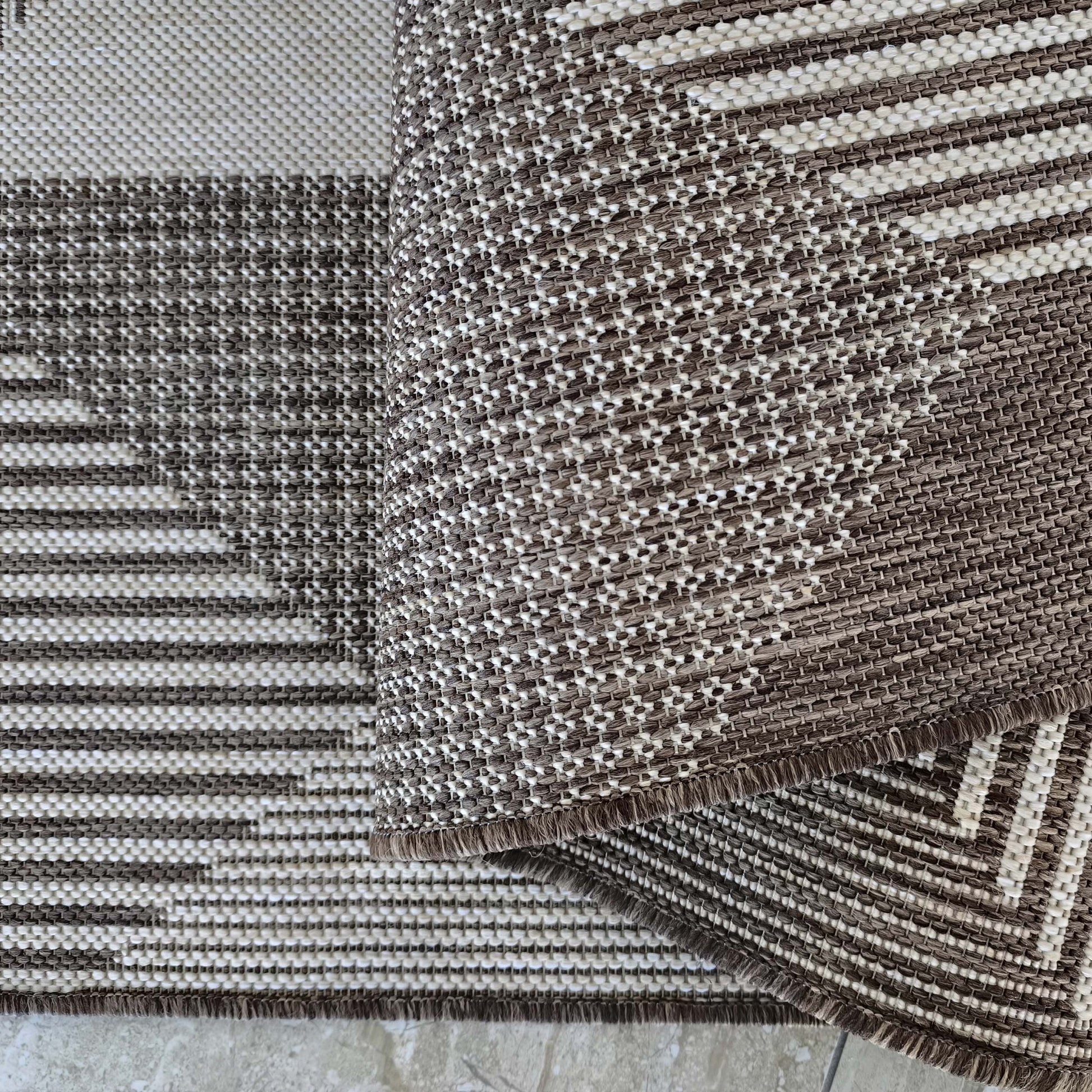 Tapis tissé marron et crème avec des motifs géométriques et des franges, partiellement replié sur un carrelage clair