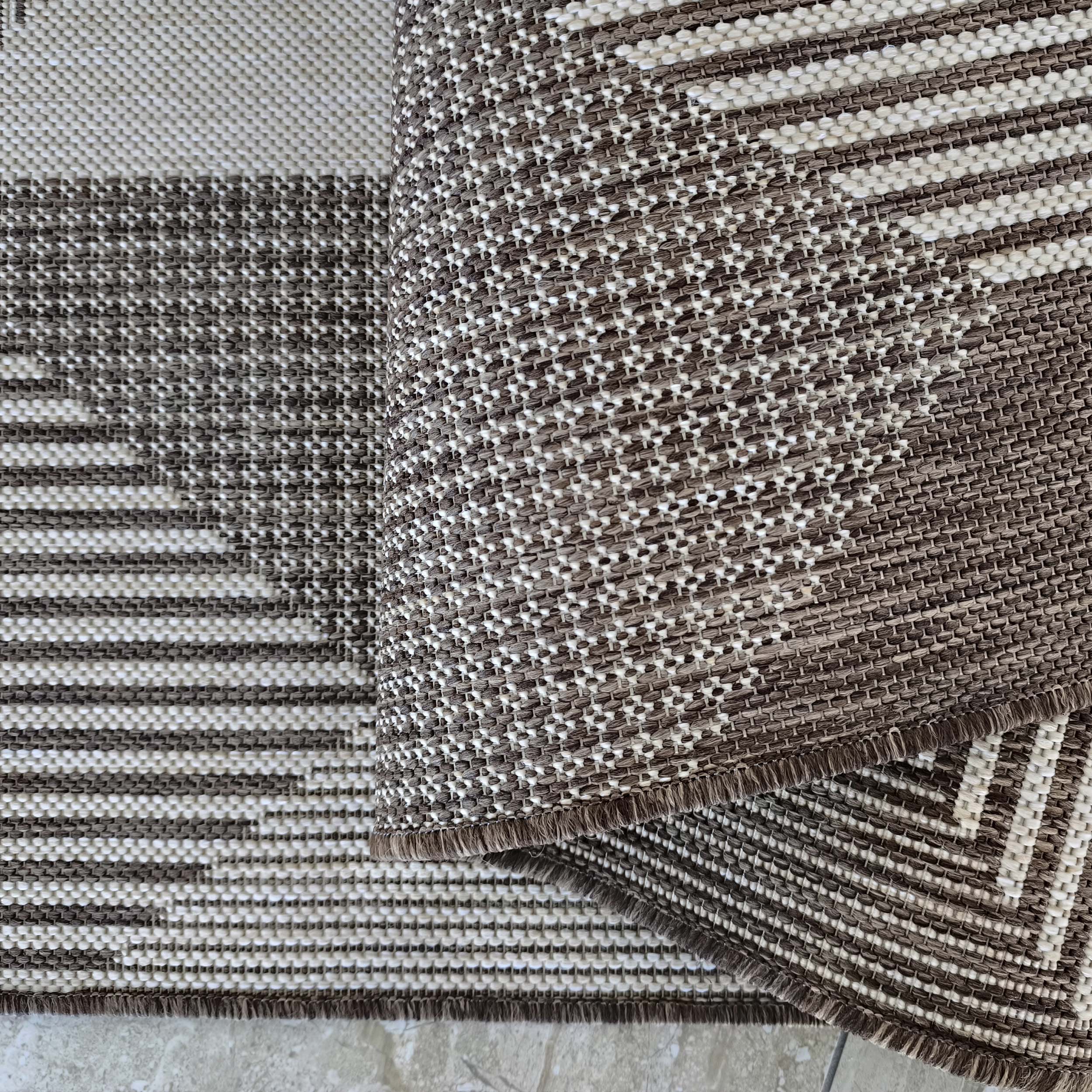 Tapis tissé marron et crème avec des motifs géométriques et des franges, partiellement replié sur un carrelage clair