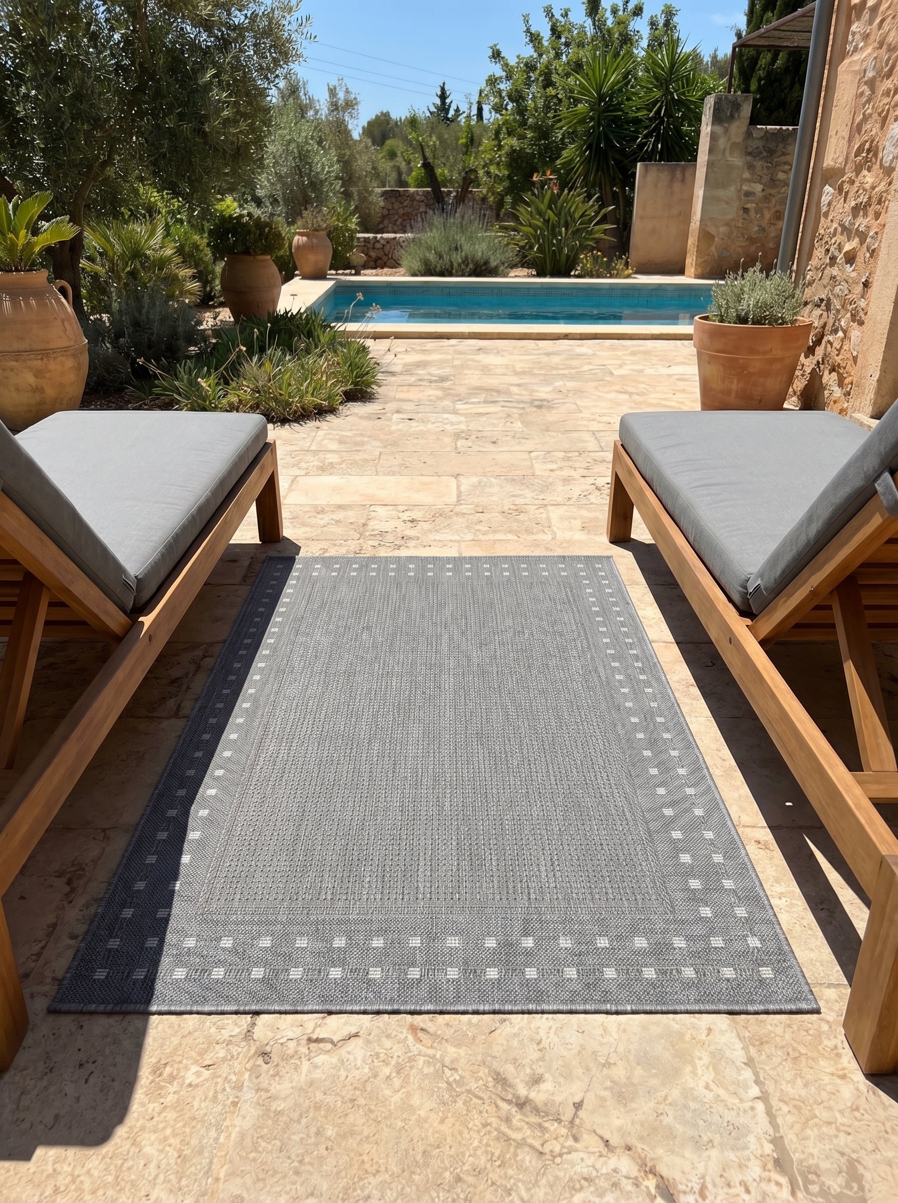 Tapis rectangulaire gris chiné, bordé d'une frise de petits carrés clairs, sur une terrasse ensoleillée