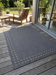 Tapis rectangulaire gris anthracite, texture tissée, avec motifs géométriques de carrés clairs en bordure, sur une terrasse en bois
