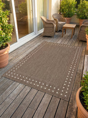 Tapis rectangulaire tissé marron-taupe, bordé de motifs de petits carrés crème, sur une terrasse extérieure en bois
