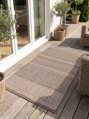 Tapis rectangulaire brun et écru aux motifs géométriques variés (chevrons, carrés), sur une terrasse en bois