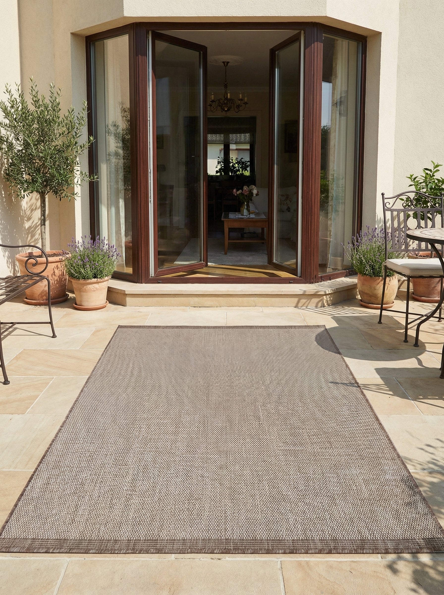 Tapis rectangulaire en tissage texturé gris-beige à bordure marron, posé sur une terrasse ensoleillée devant une maison