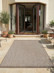 Tapis rectangulaire en tissage texturé gris-beige à bordure marron, posé sur une terrasse ensoleillée devant une maison