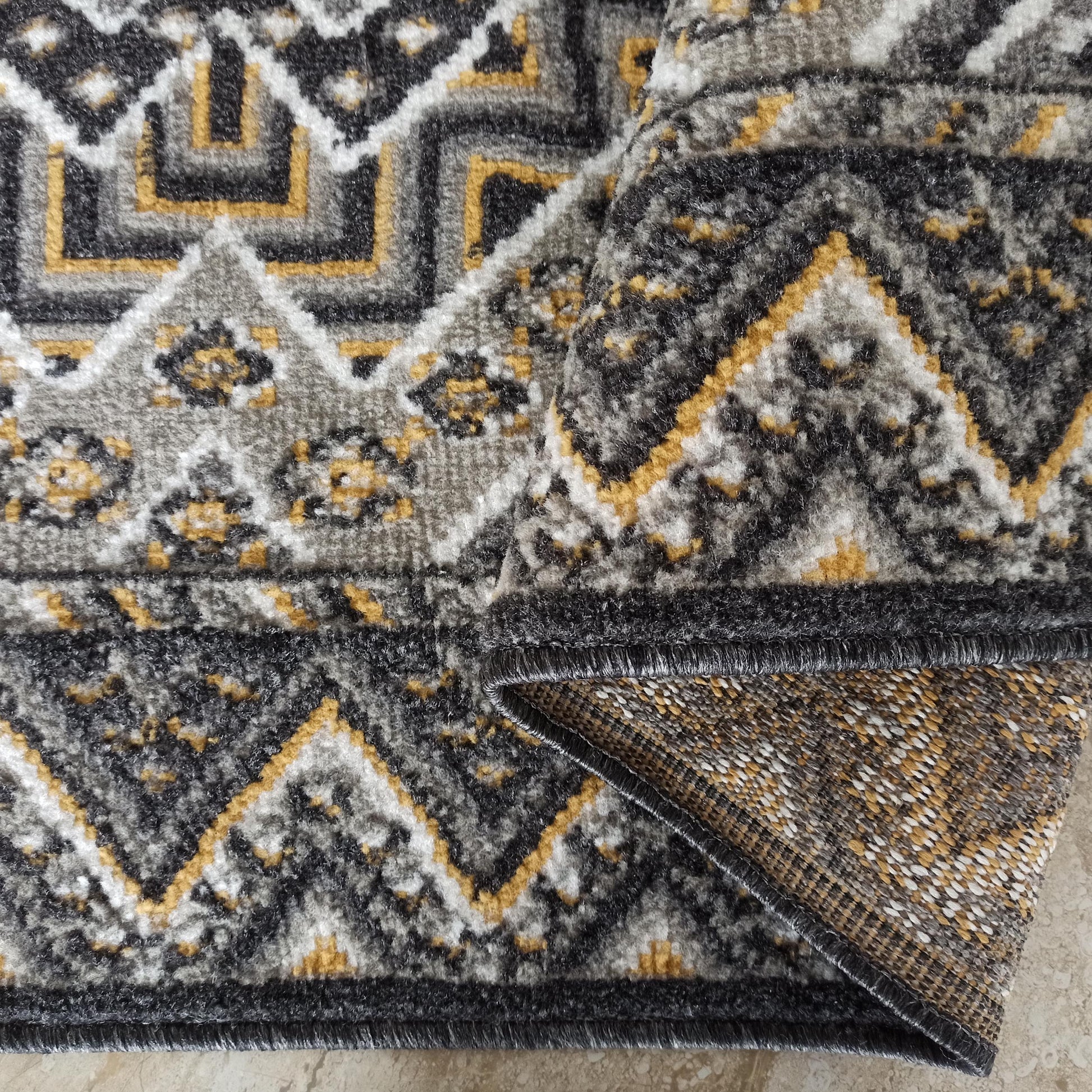 Tapis à motifs géométriques gris, noir, blanc et jaune ocre. Le coin replié révèle l'envers tissé