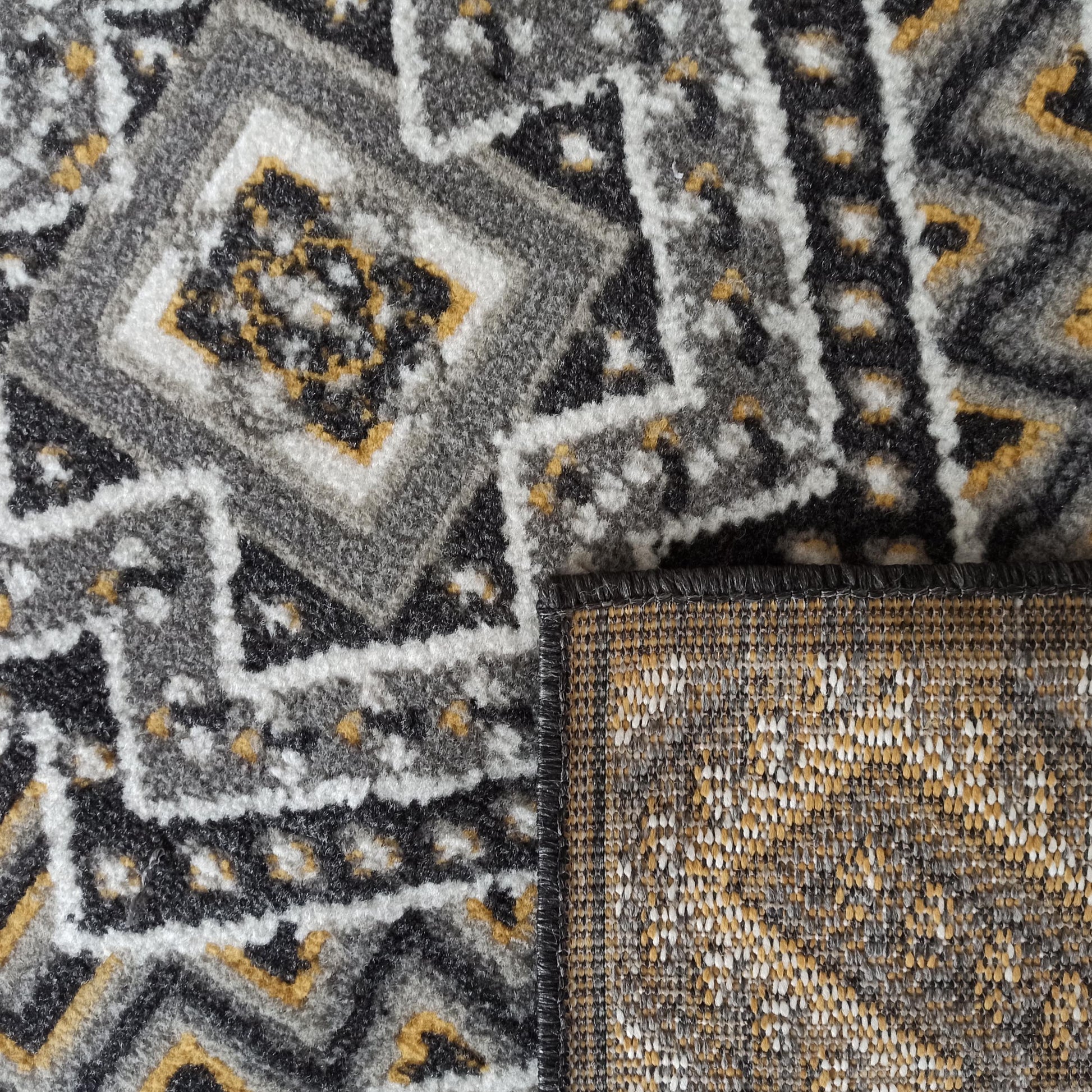 Tapis aux motifs géométriques tissés gris, noir, blanc et jaune ocre. Un coin est relevé pour montrer l'envers tressé