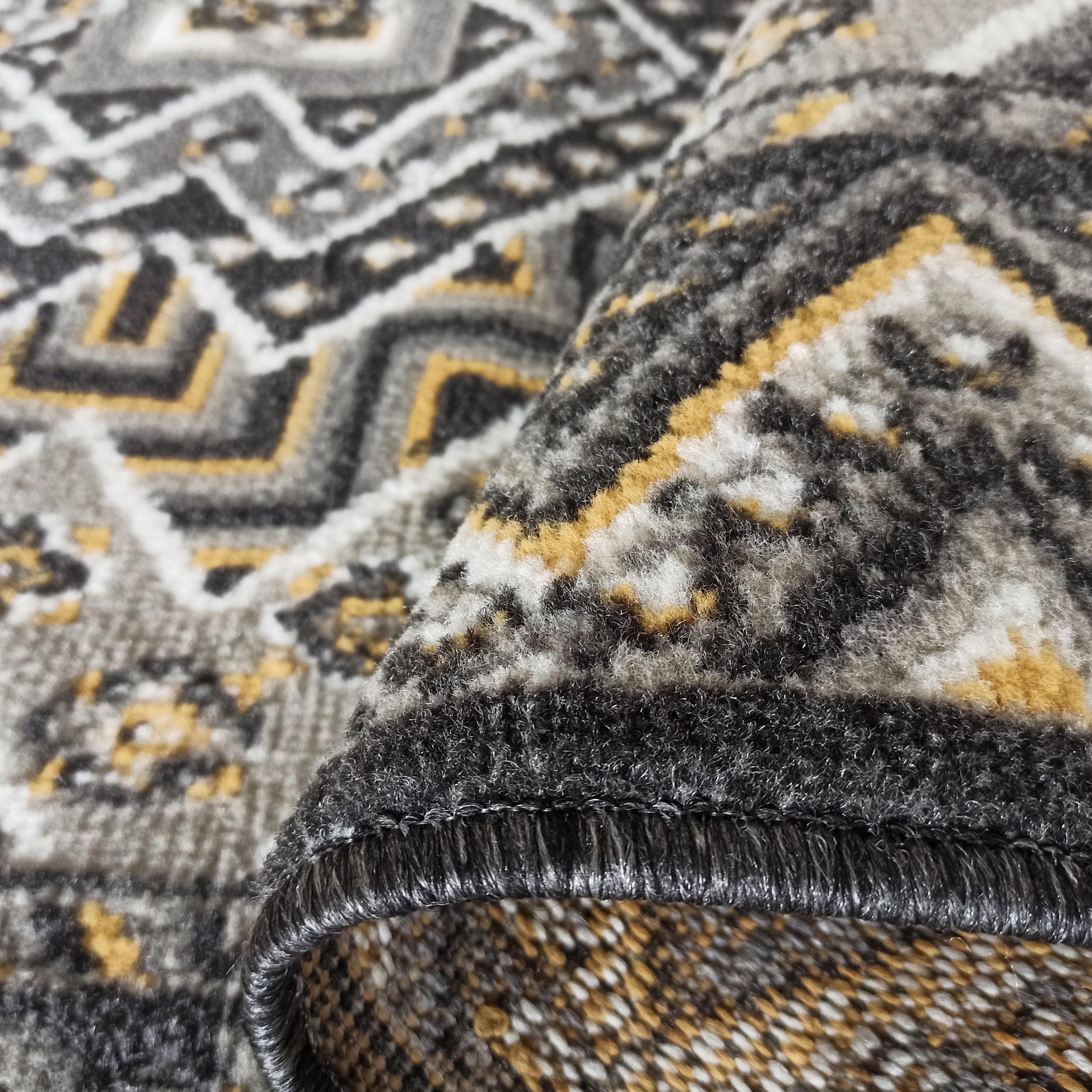 Tapis doux aux motifs géométriques gris, noirs, blancs et jaunes, avec un bord roulé montrant sa texture poilue